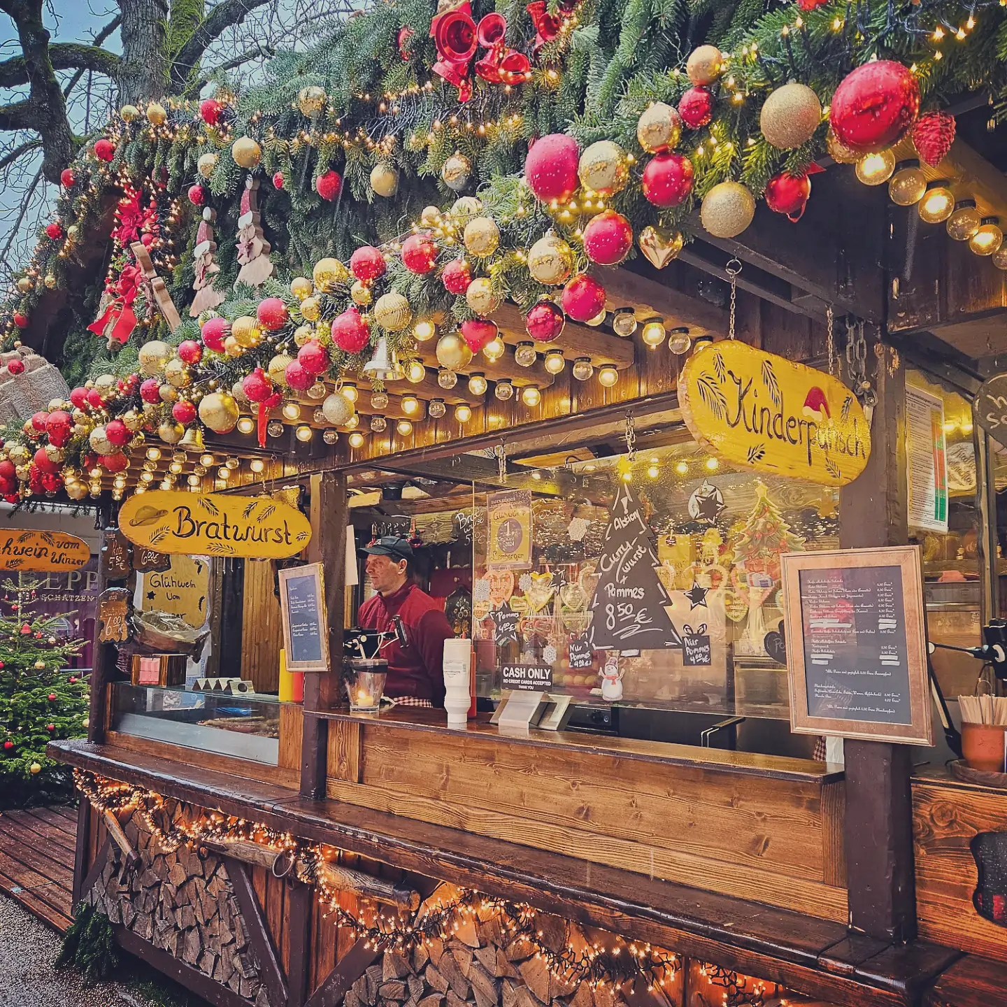 Wooden stall selling Bratwurst and Kinderpunsch, decorated with garlands and glowing red and gold ornaments.
