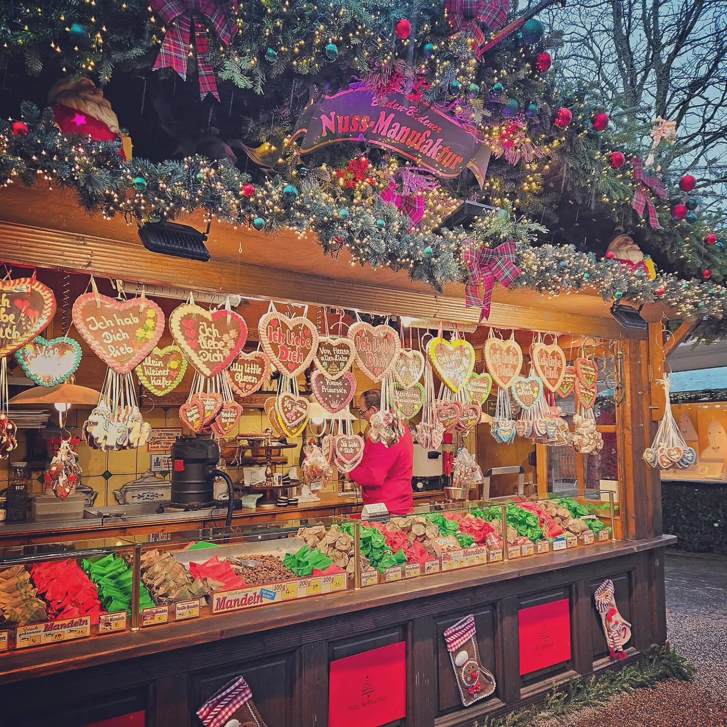 Christmas market stall selling gingerbread hearts and candied nuts, decorated with greenery and colorful ornaments.