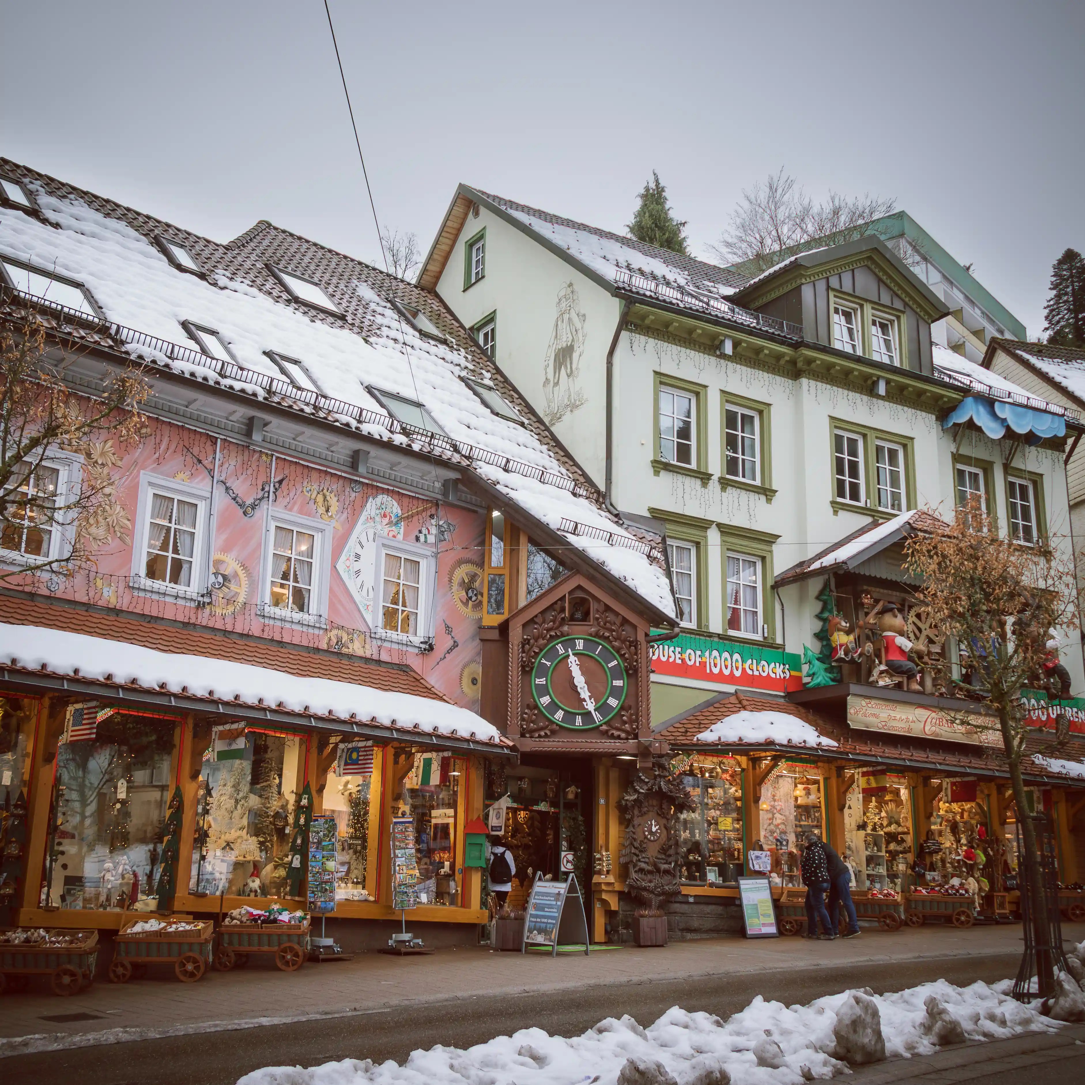 Street view of colorful buildings in Triberg with mural-painted façades and shop signs reading “House of 1000 Clocks.”