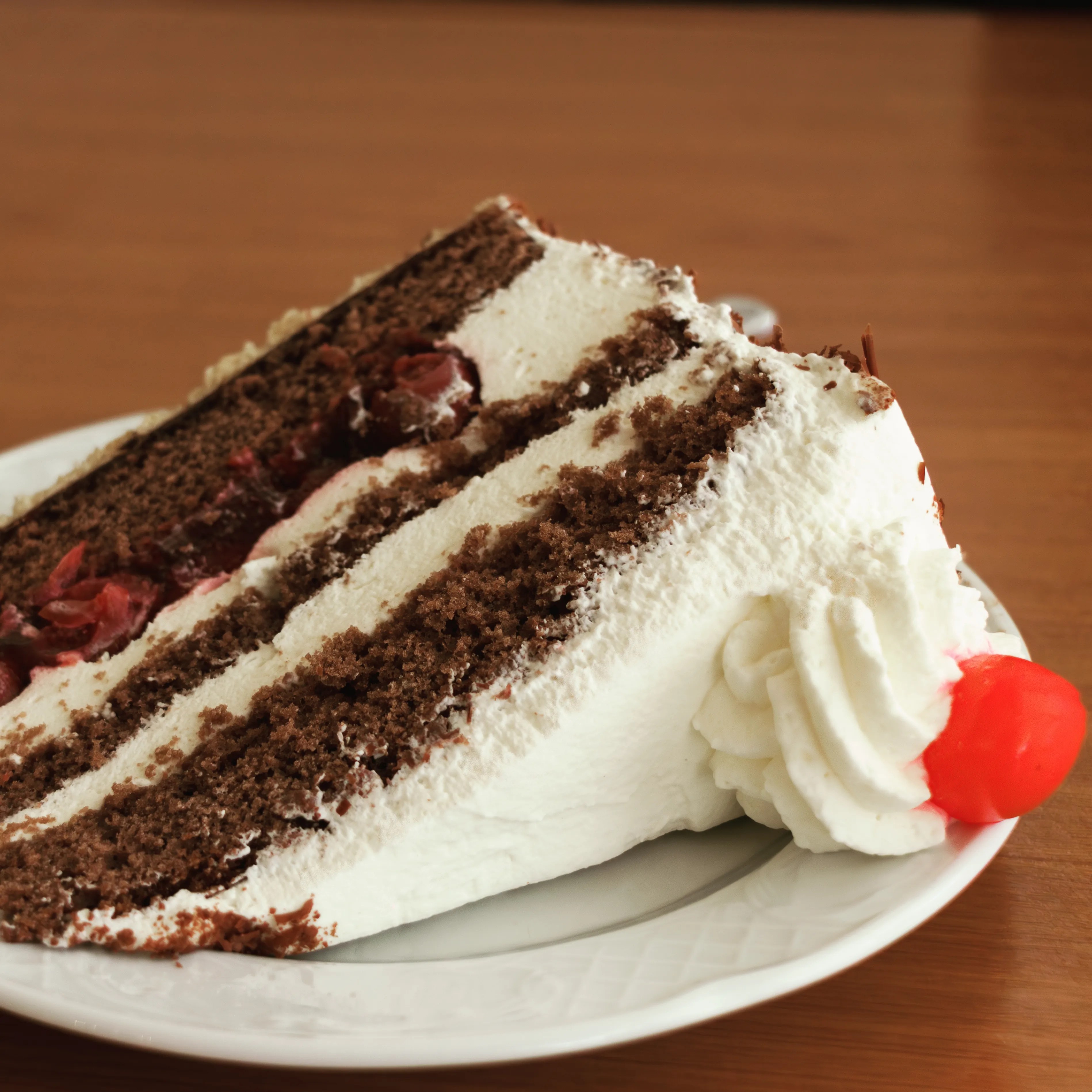 Close-up of a slice of Black Forest cake with layers of chocolate sponge, whipped cream, cherries, and a maraschino cherry on top.