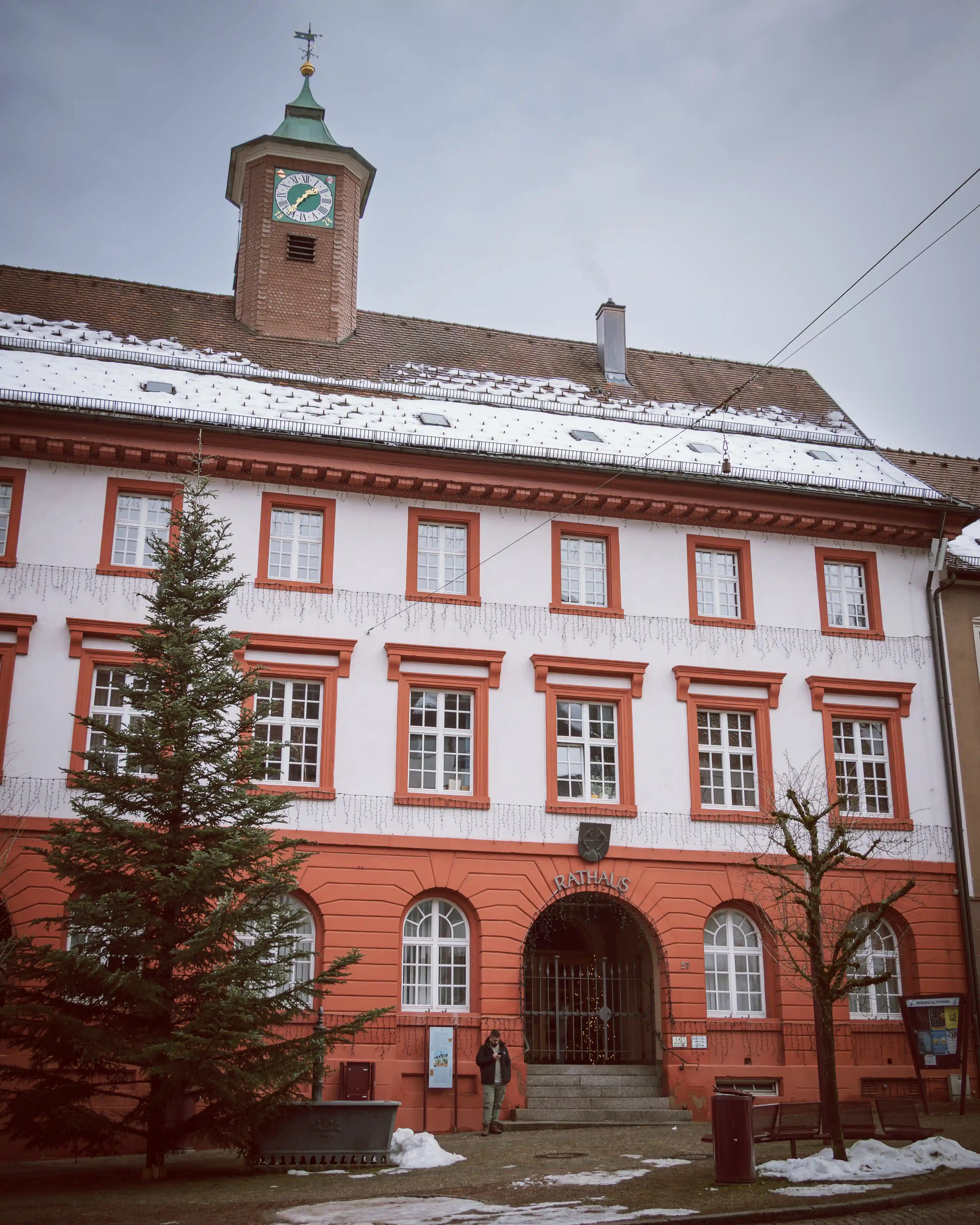 Red and white facade of the Triberg Rathaus (town hall) with a clock tower and Christmas lights above the entrance.