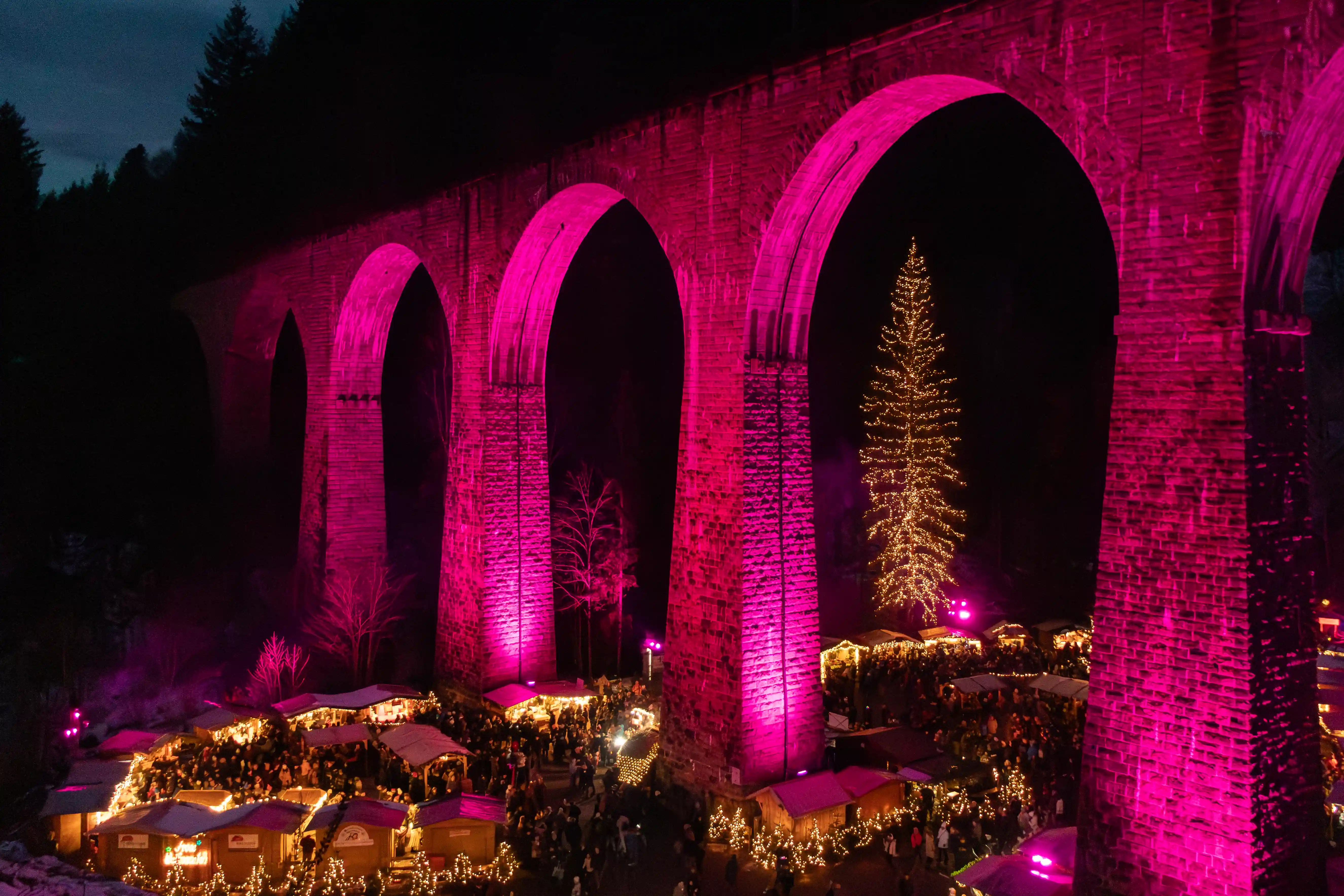 Night view of the Ravenna Gorge Christmas Market with bright pink lighting on the viaduct and a golden Christmas tree at the center.