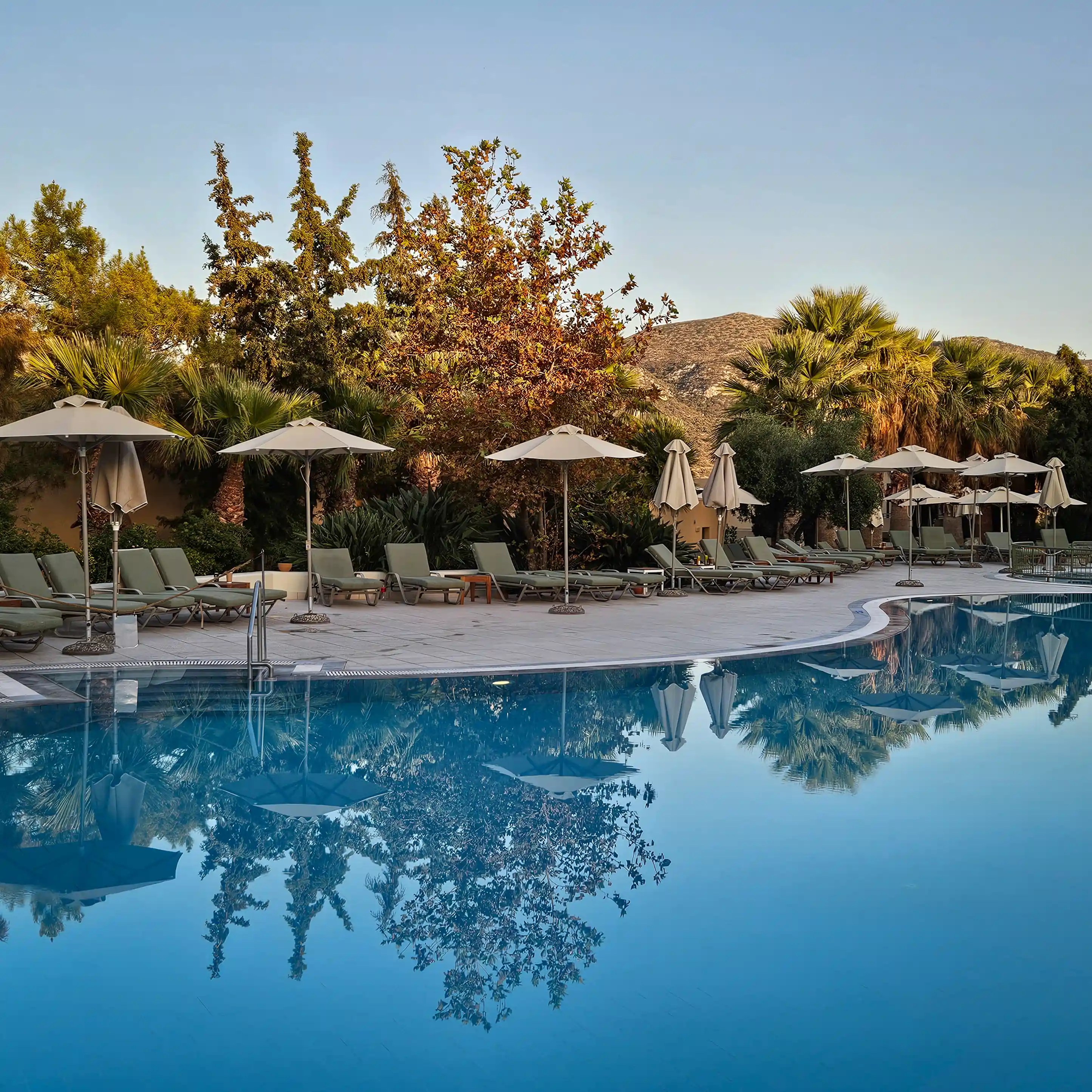 Calm resort swimming pool at sunrise with palm trees and sun loungers, Village Heights Resort in Hersonissos, Crete.