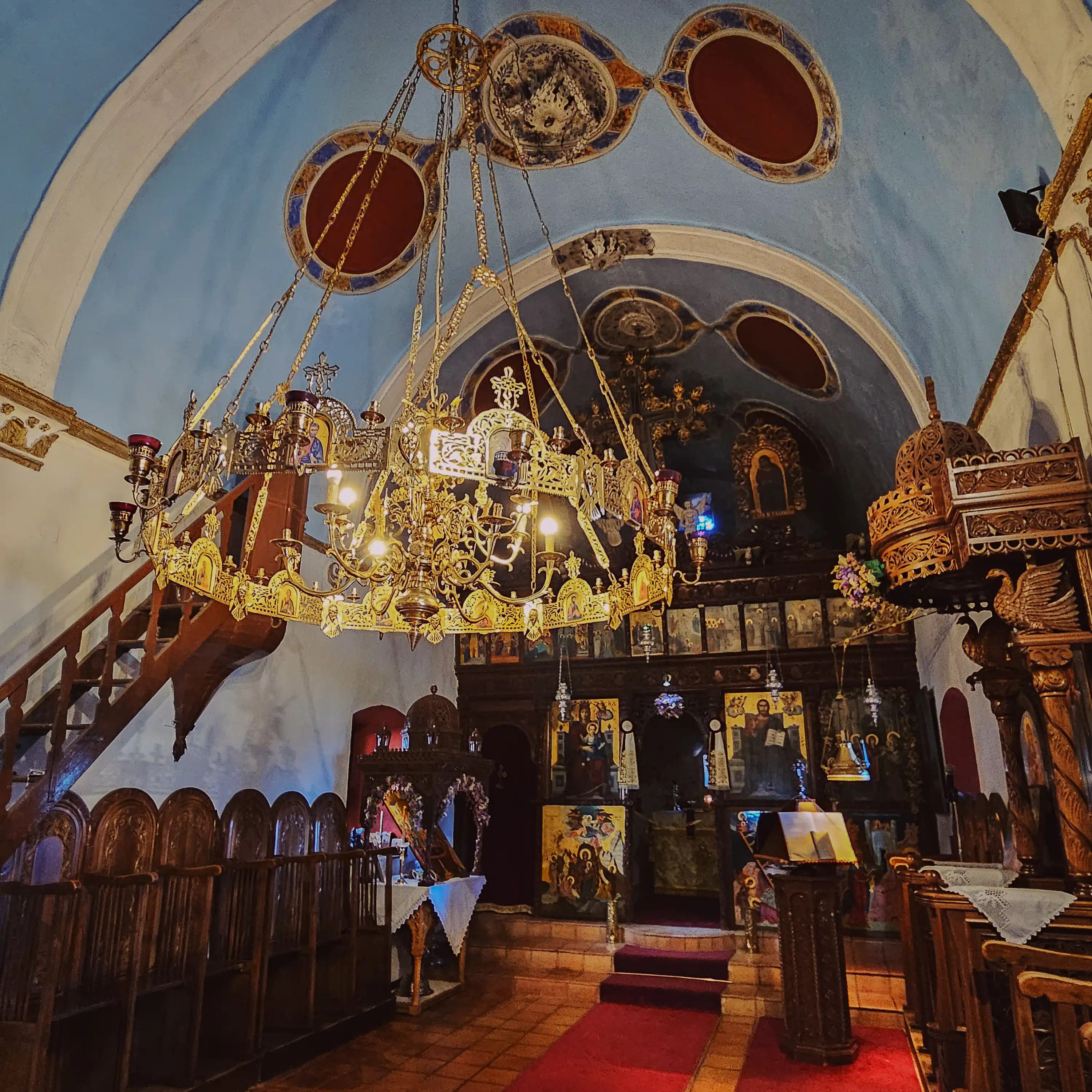 Interior of the Holy Monastery of Vidiani with an ornate gold chandelier, religious paintings, red carpet, and carved wooden seating.