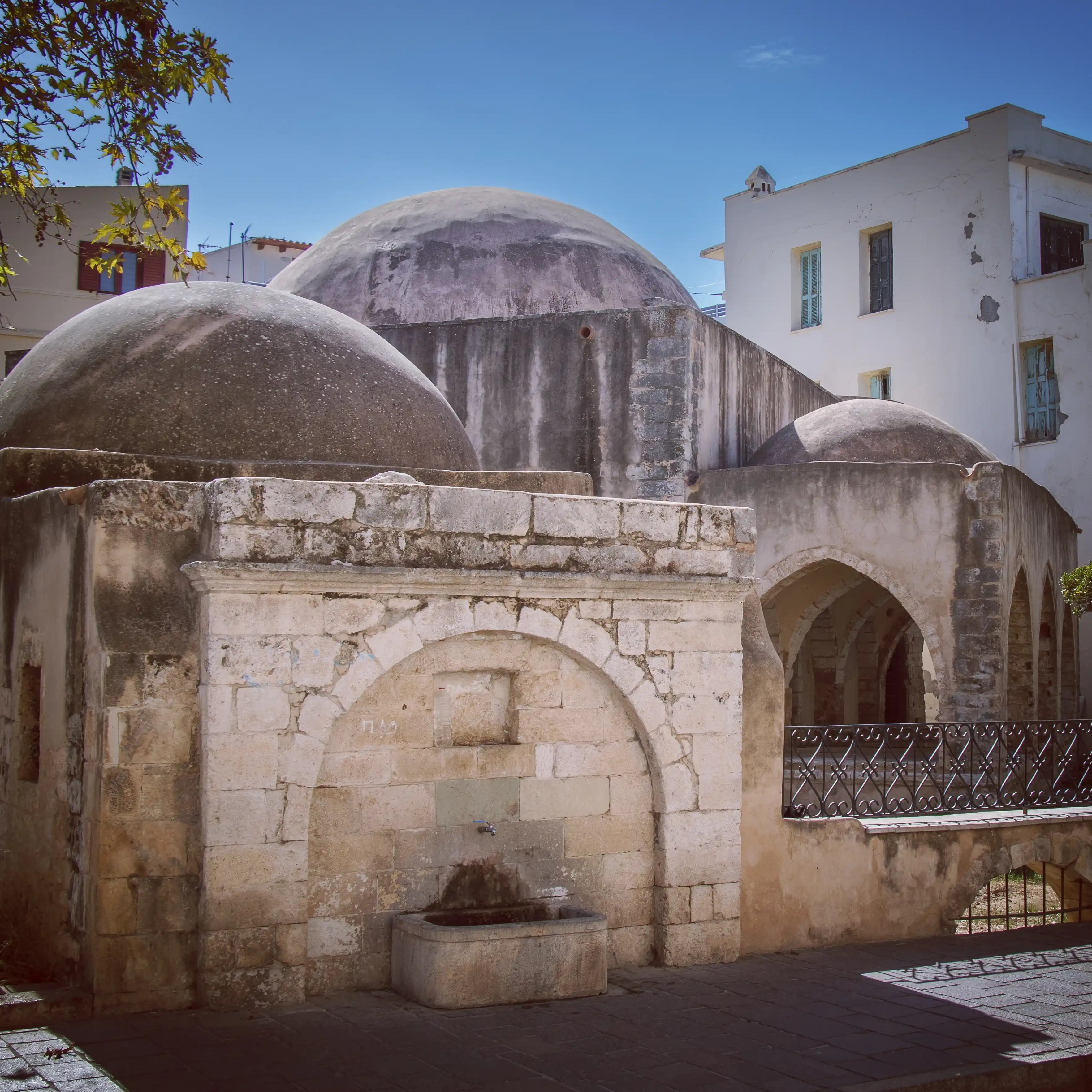 The domed exterior of the Kara Musa Pasha Mosque in Rethymno with a stone fountain in front.
