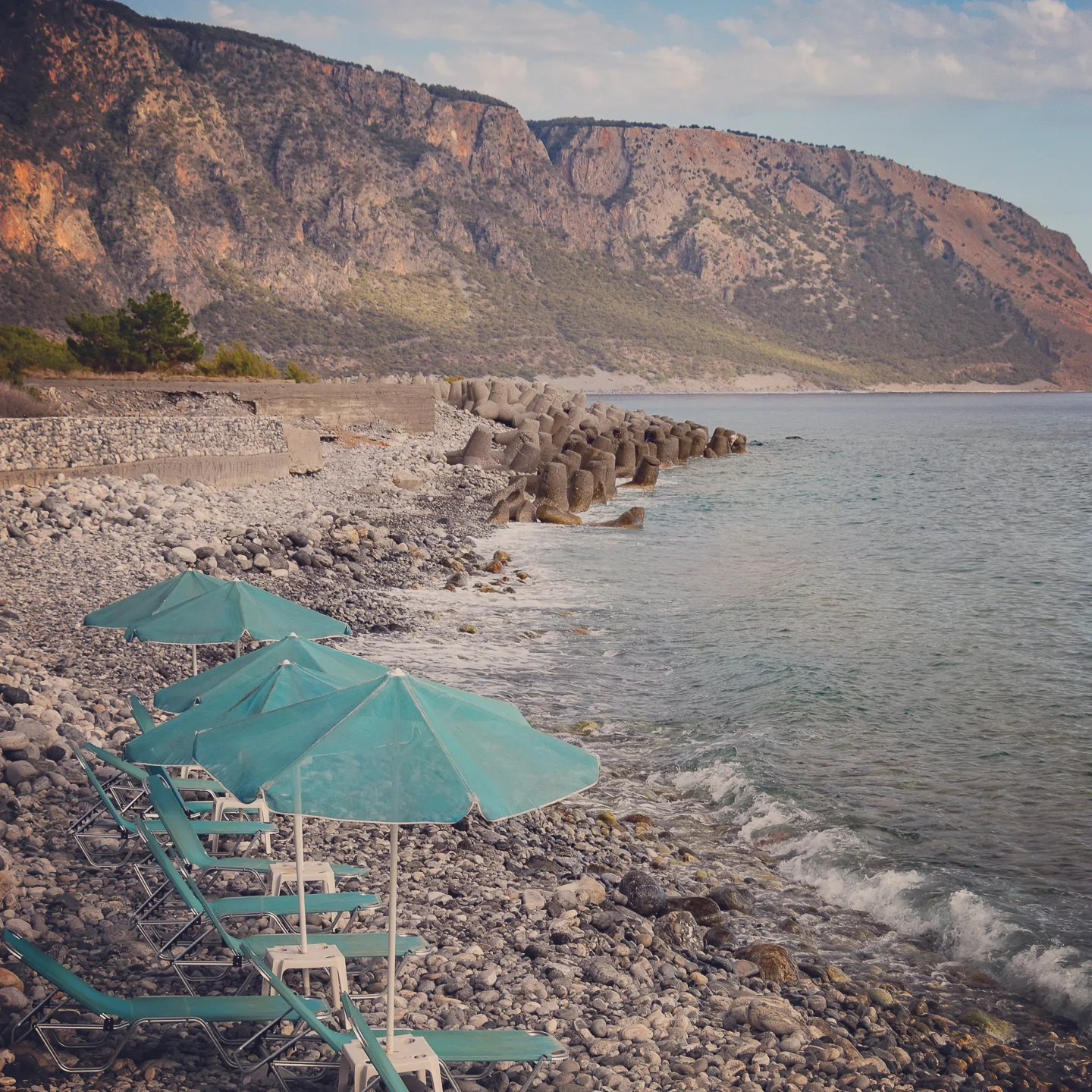 Row of turquoise beach umbrellas on a rocky shoreline with mountains along the coast.