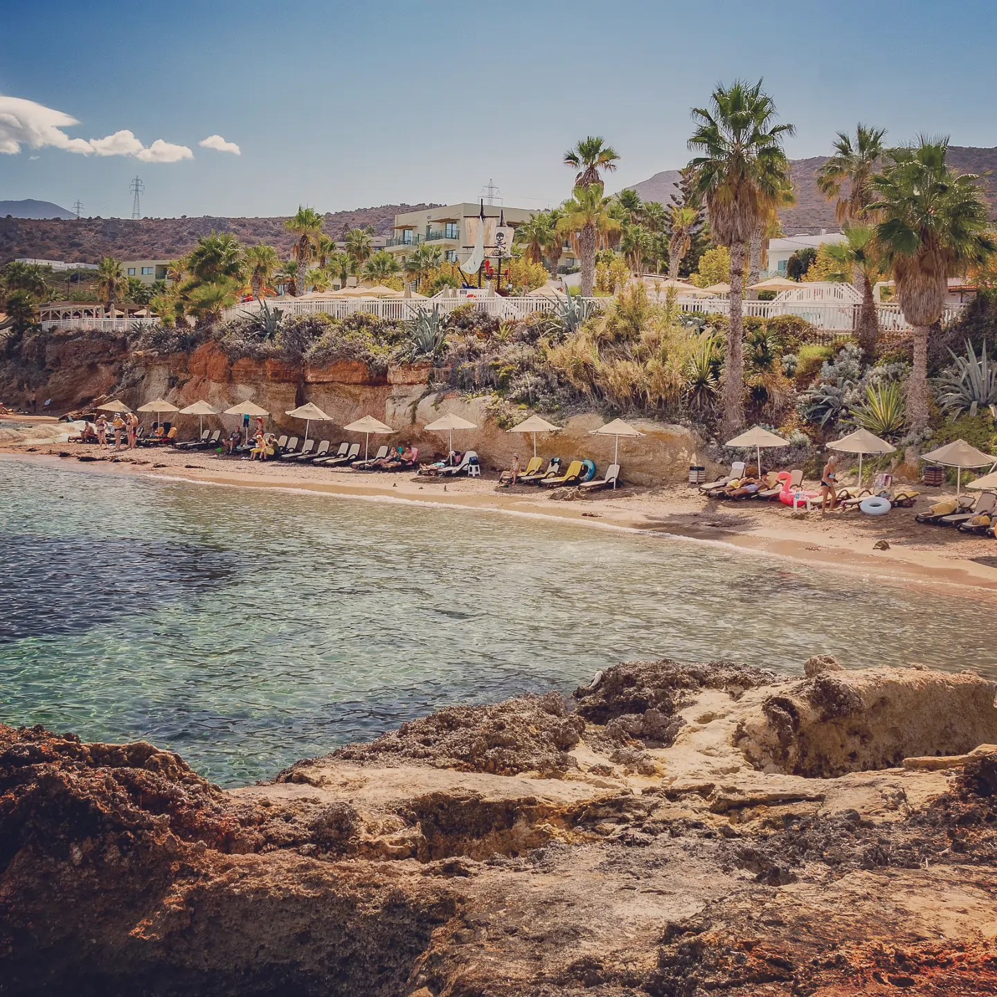 Sandy cove with sunbeds, umbrellas, and palm trees along the Hersonissos coast.
