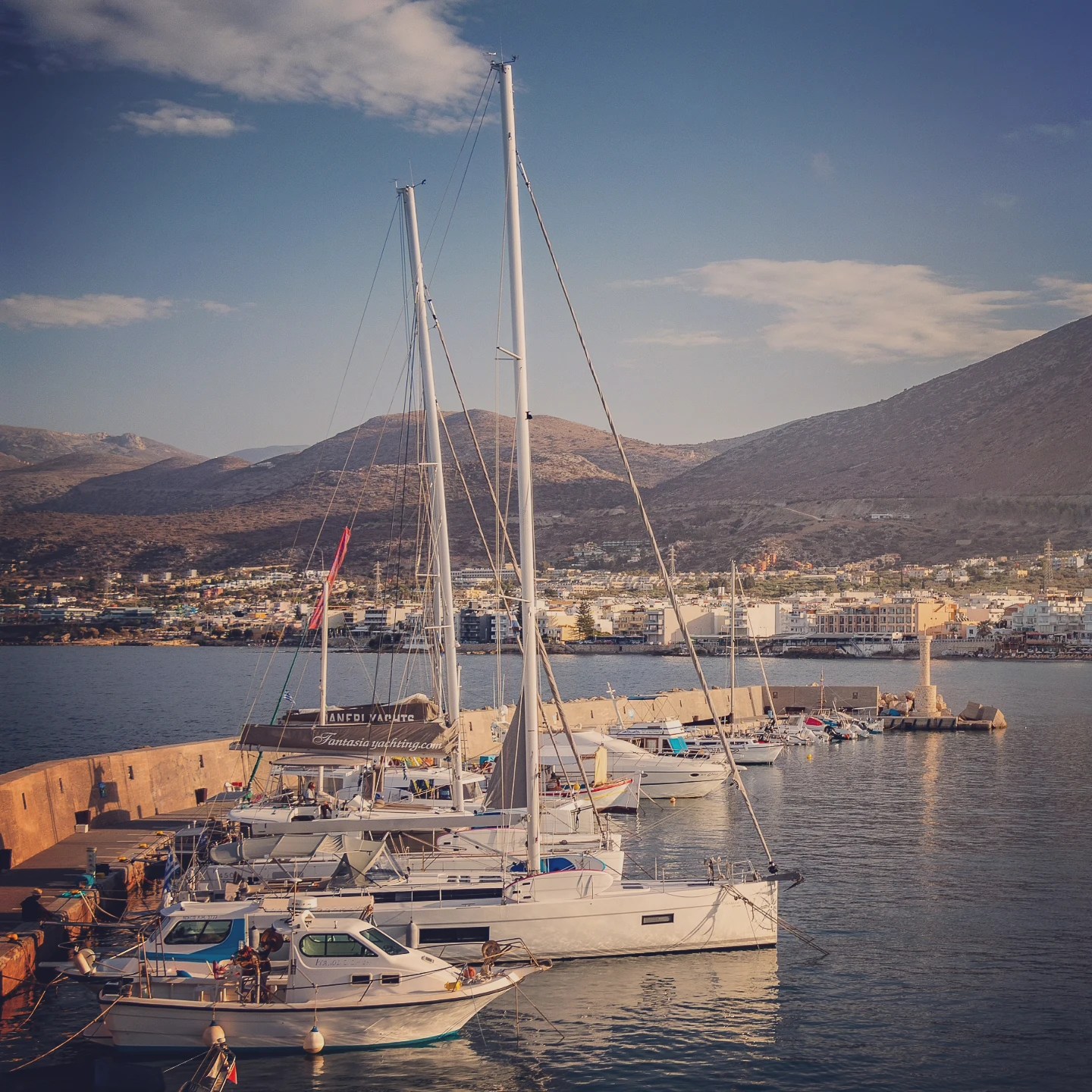 Sailboats docked at Hersonissos Harbor with mountains and town buildings in the background.