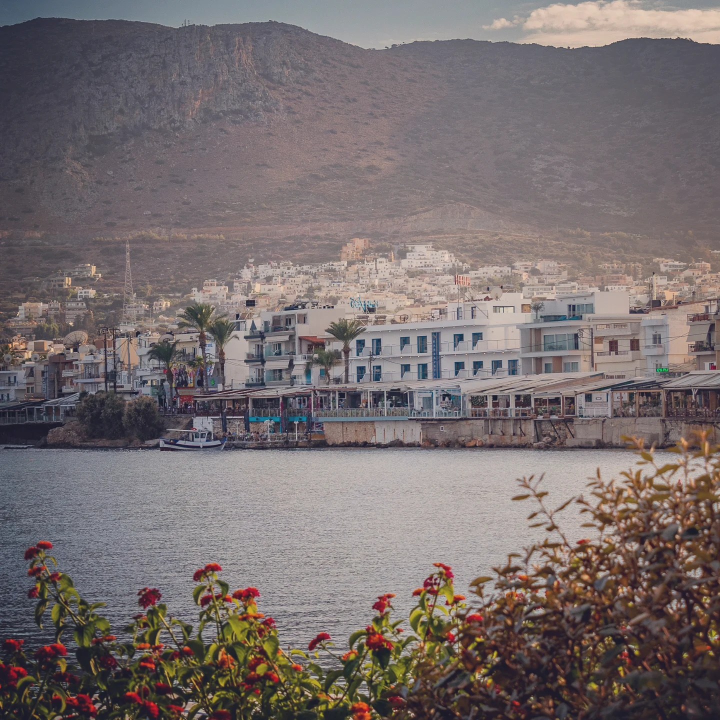 View of Hersonissos waterfront with hotels and homes along the bay, backed by rugged mountains.