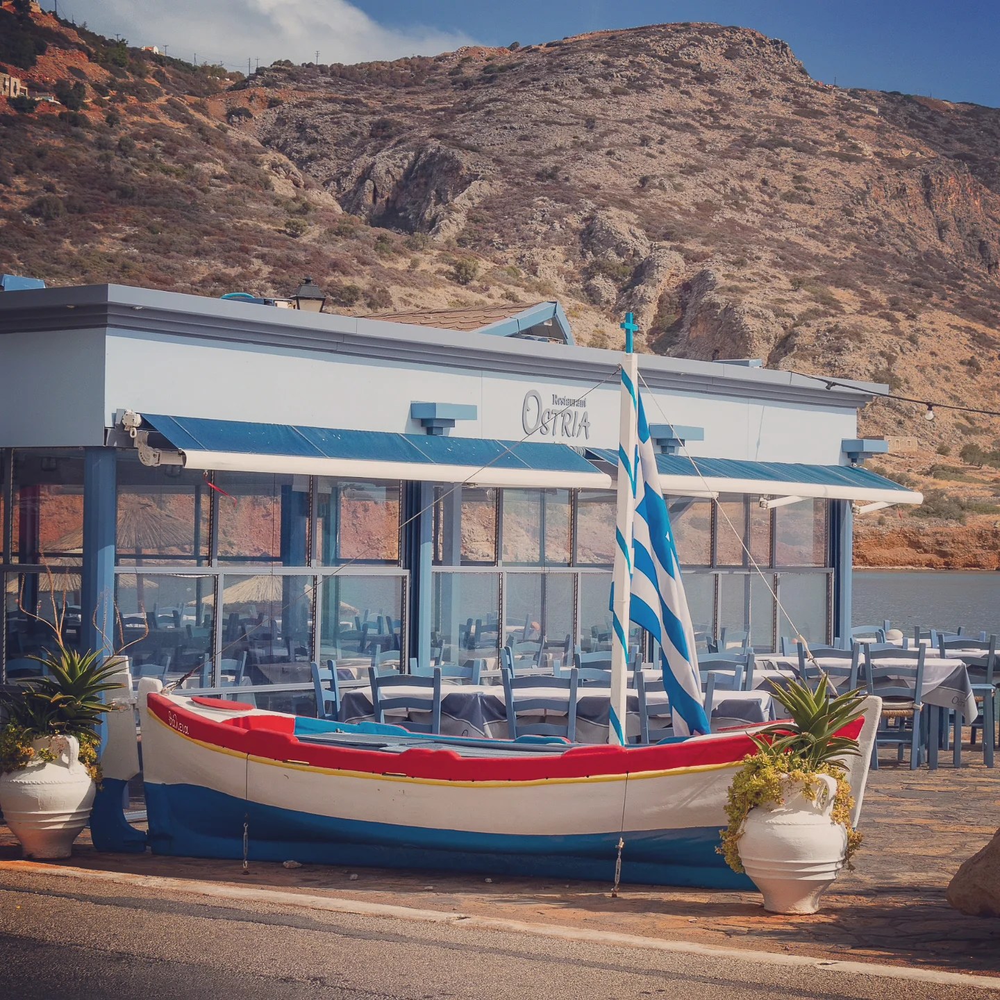 A small traditional boat sits in front of Restaurant Ostria with the rocky hills of Plaka rising behind it.