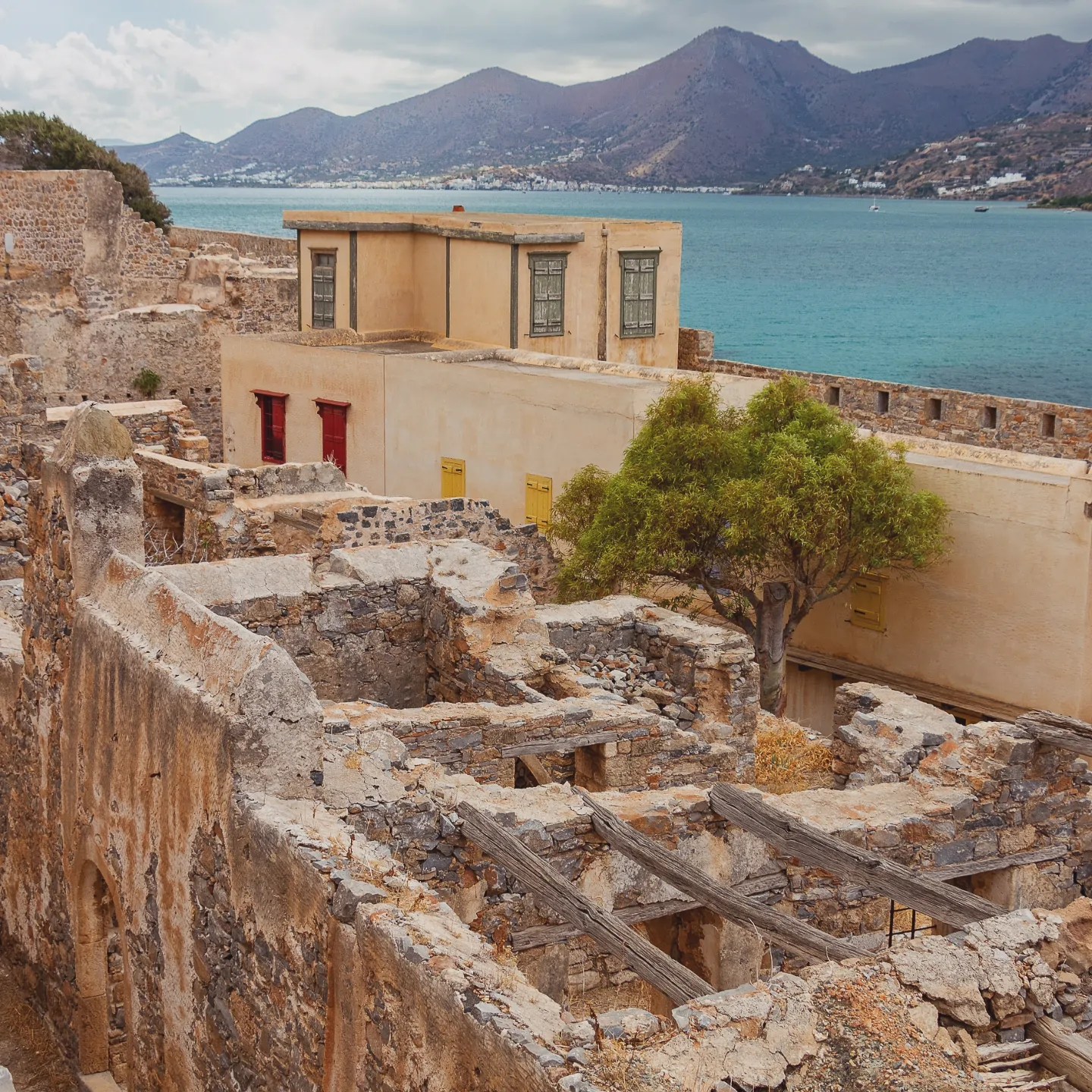 A view over Spinalonga’s stone ruins toward a pale yellow building and the turquoise bay beyond.