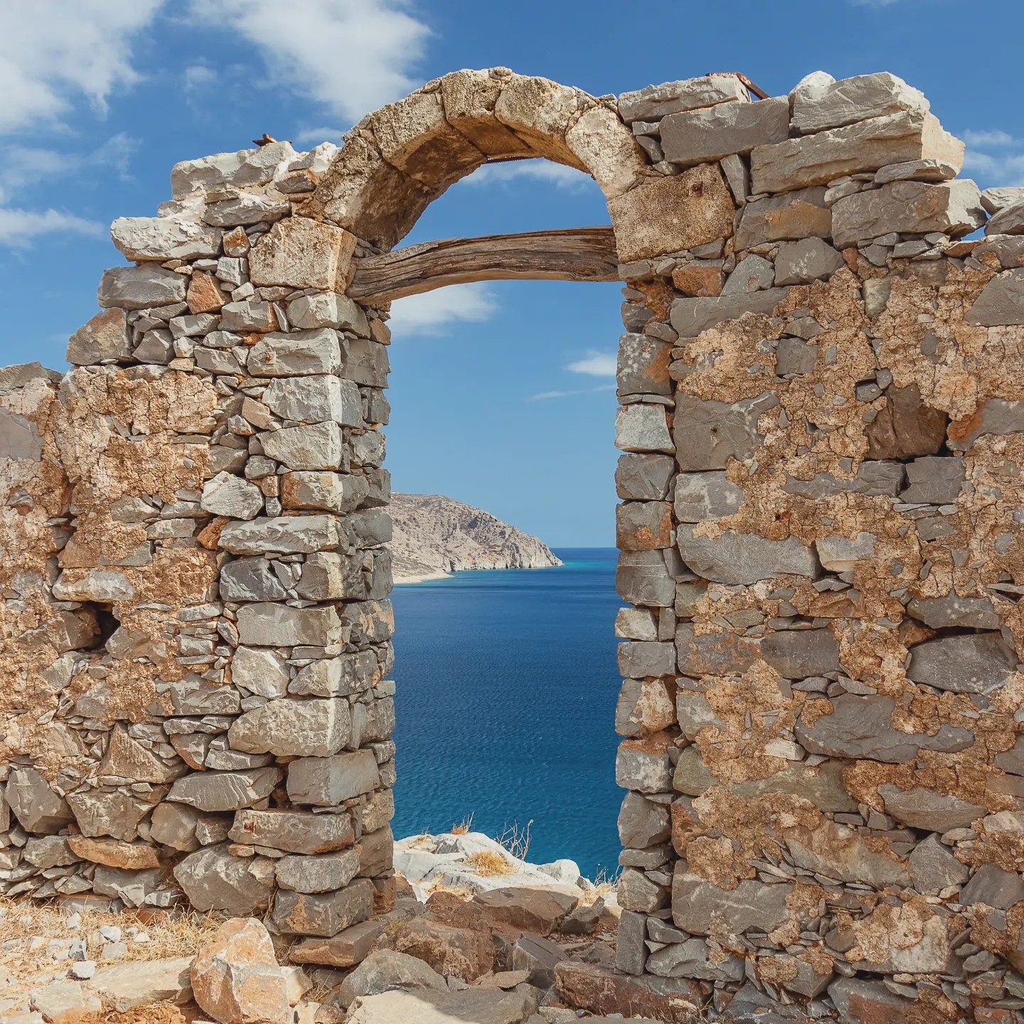 A stone archway in a ruined wall on Spinalonga framing a view of the deep blue sea and nearby coastline.