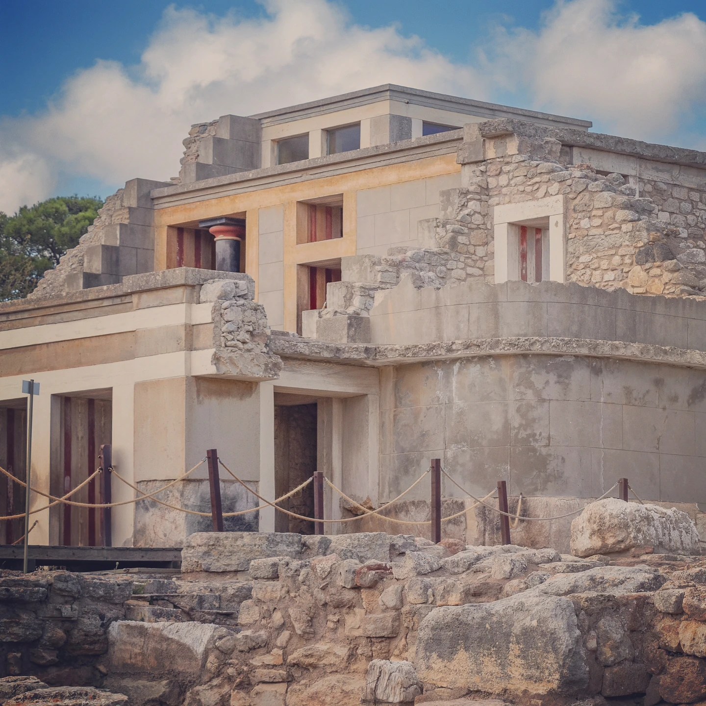 Restored multi-story structure at the Palace of Knossos with stone walls, red columns, and ochre trim under a blue sky.