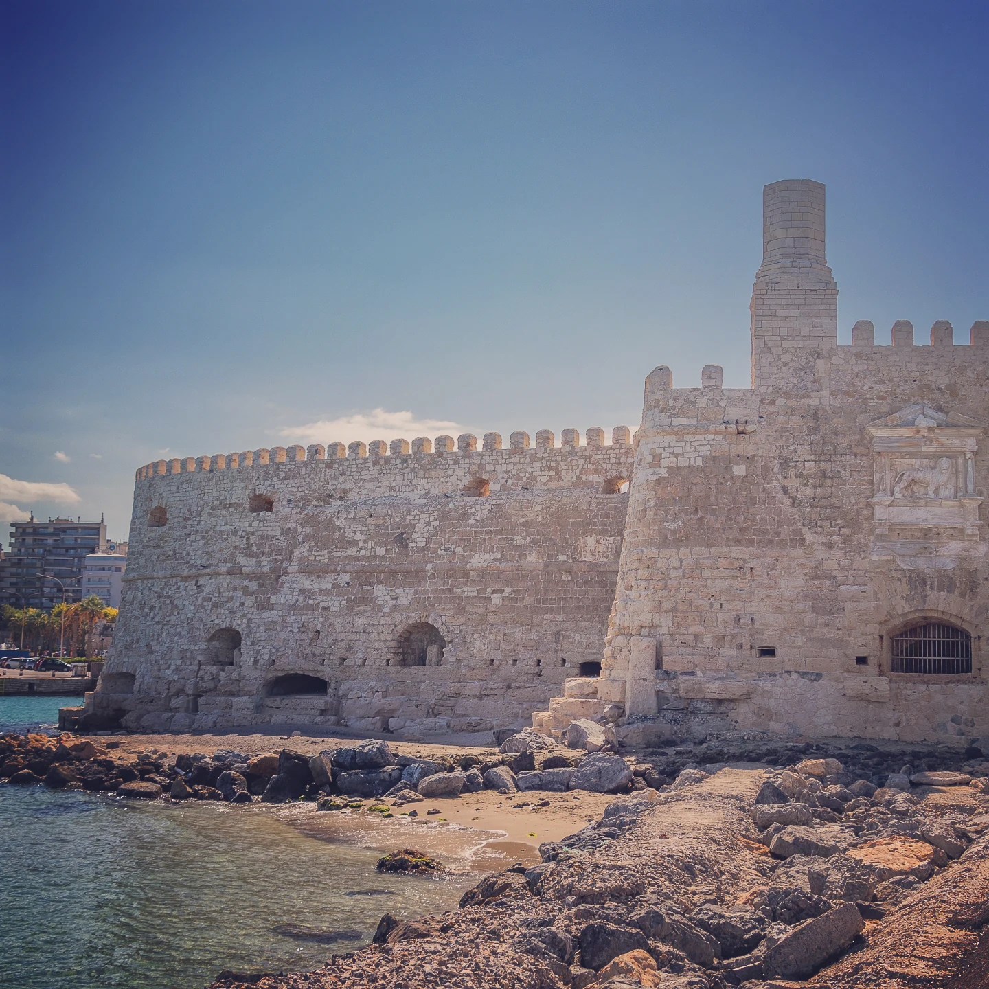 Koules Fortress seen from the shoreline with waves and rocks in the foreground.