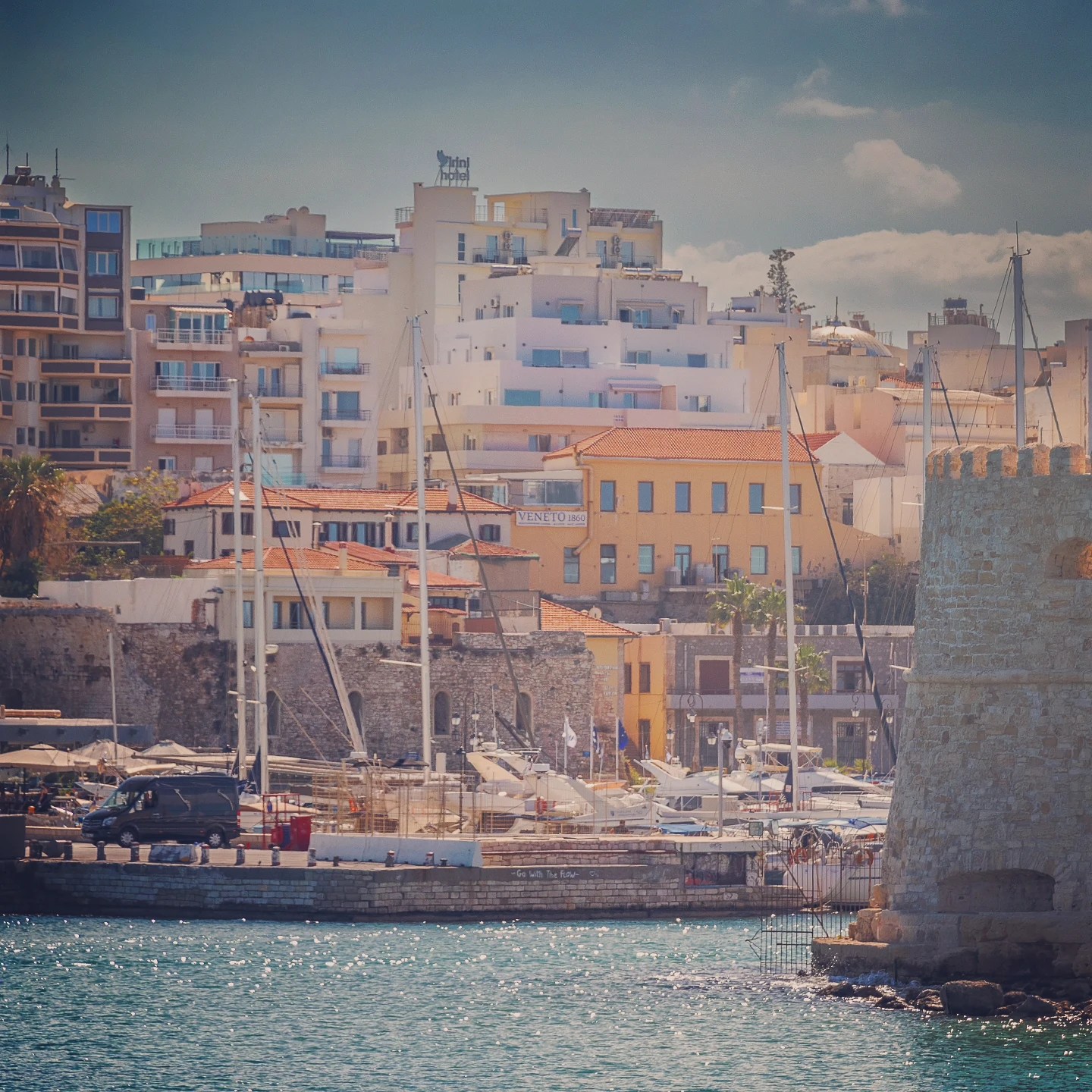 View of Heraklion’s marina with sailboats, colorful buildings, and part of the Venetian harbor walls.
