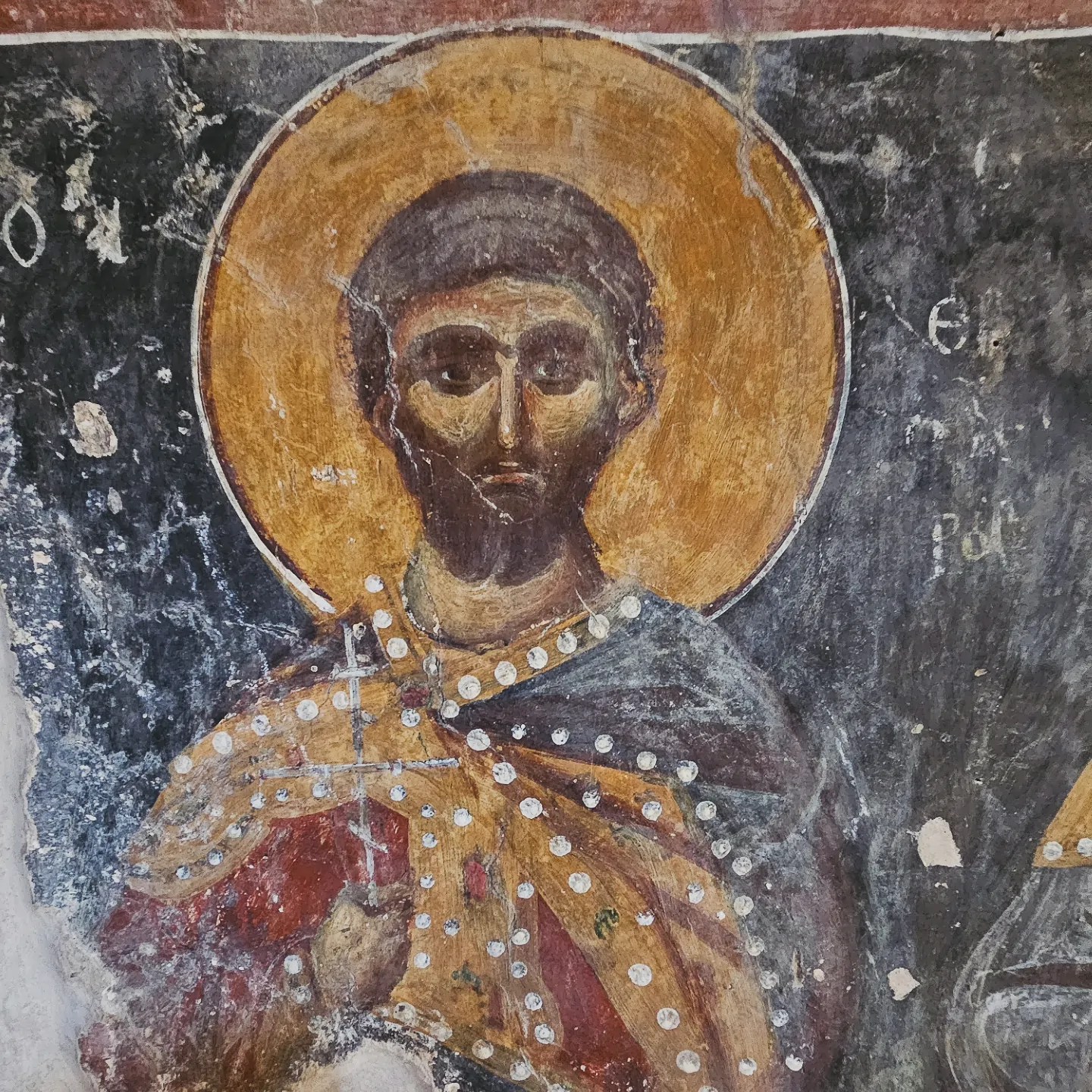 Close-up of a Byzantine fresco depicting a male saint with a dark beard, golden halo, and richly patterned robes.
