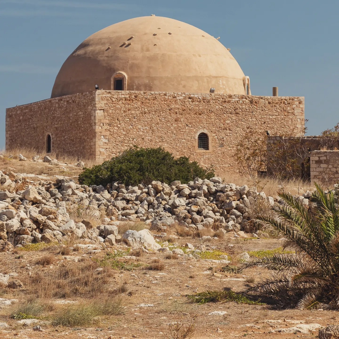 The large domed mosque inside the Rethymno Fortezza surrounded by rocky, dry ground and scattered stones.