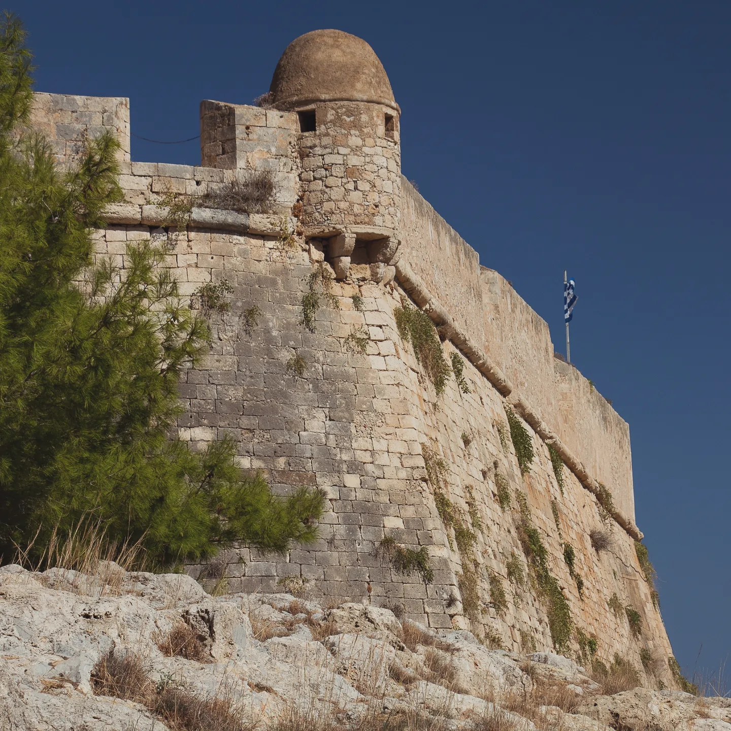 A corner tower and high stone wall of the Rethymno Fortezza rising above rocky ground with a Greek flag in the distance.