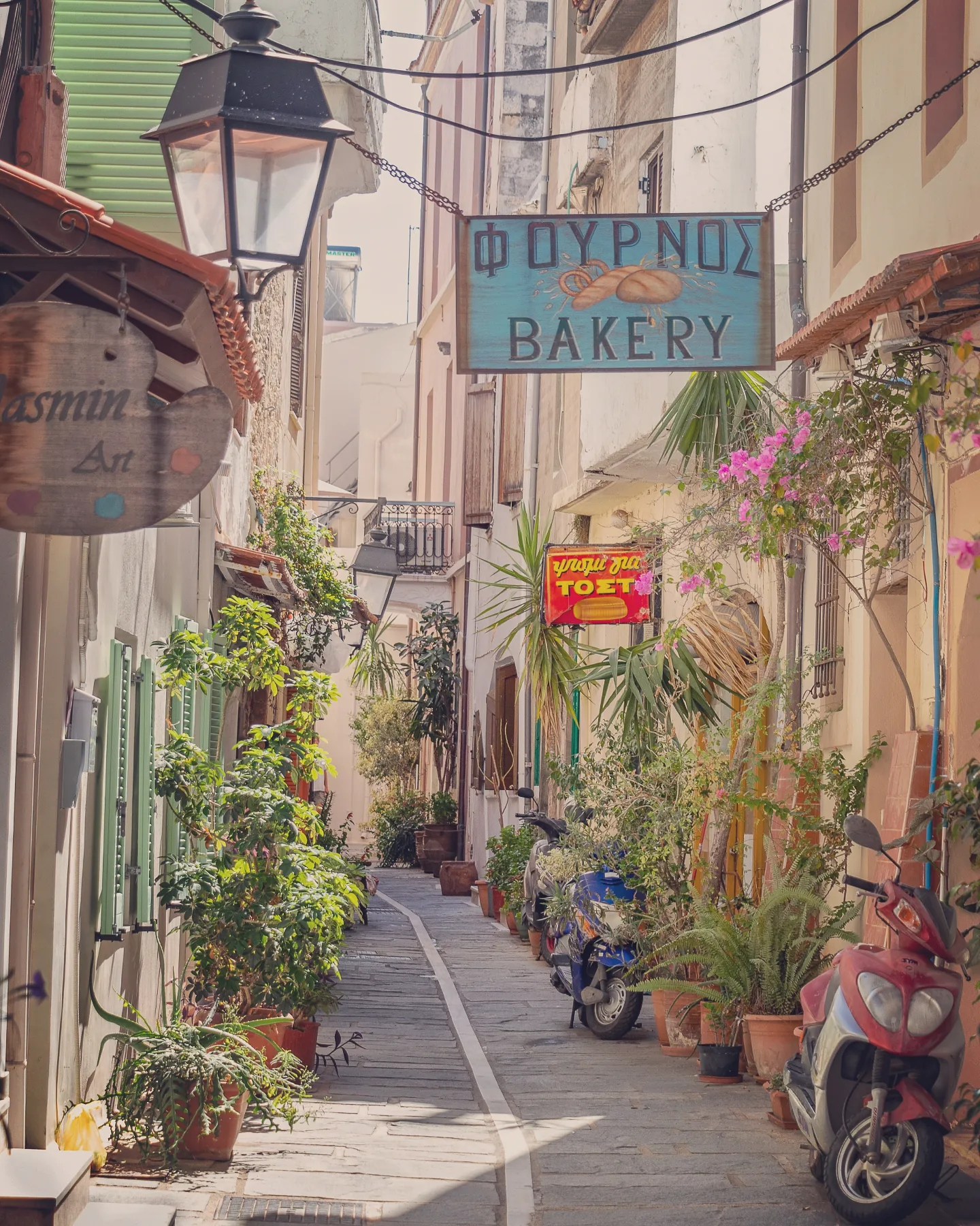 A narrow Rethymno alley lined with plants, scooters, and signs including a blue “Bakery” sign written in Greek and English.