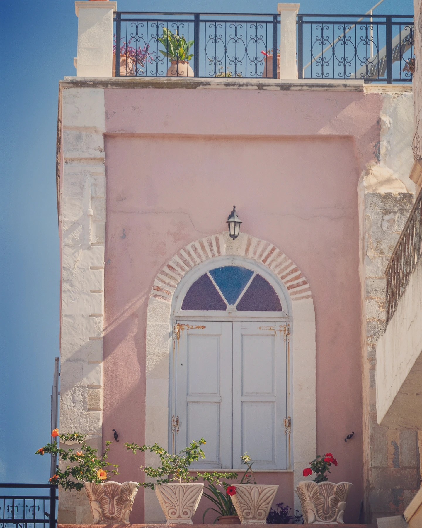 A pink building in Rethymno with a white arched window, balcony railing above, and flower pots along the ledge.
