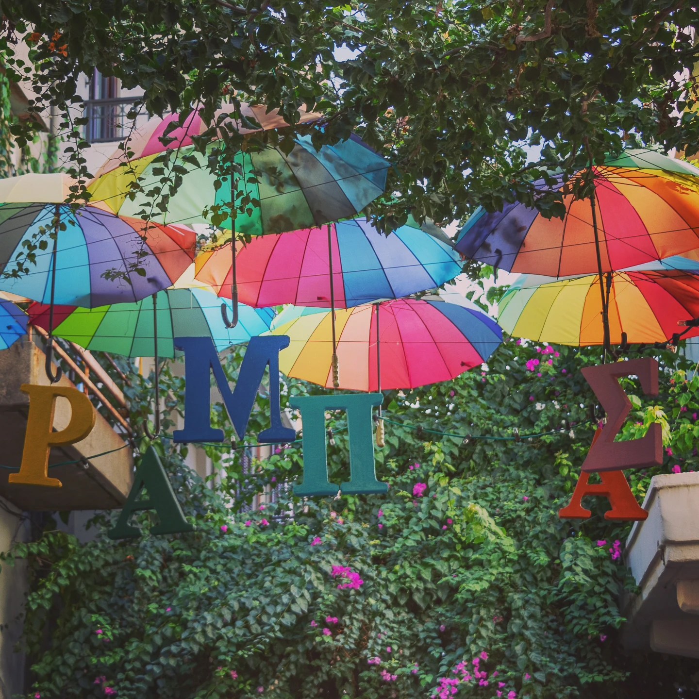 Brightly colored umbrellas hang above a narrow street with greenery and Greek letters suspended among them.