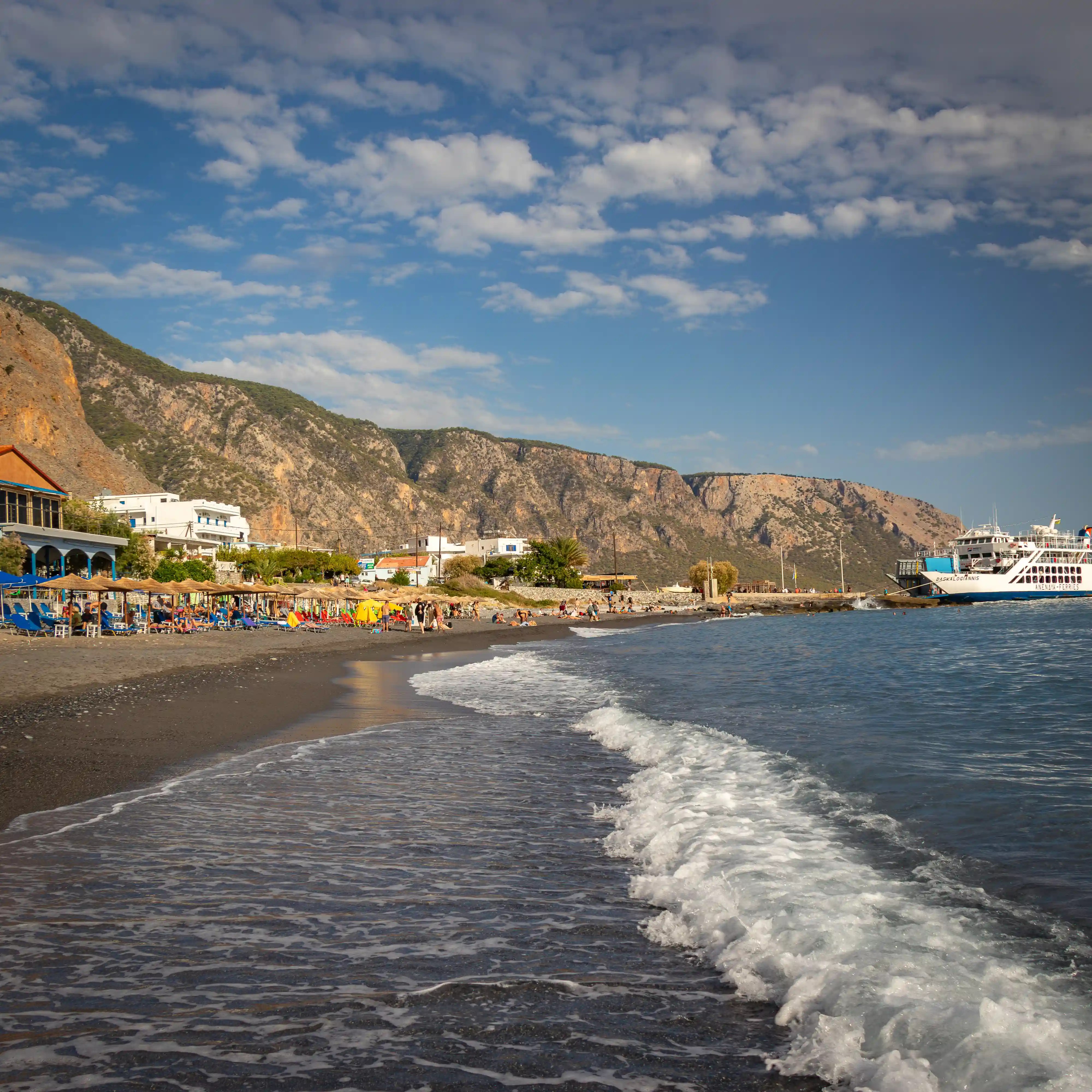 Beach at Agia Roumeli with waves, sunbathers, and a ferry docked near the shore.