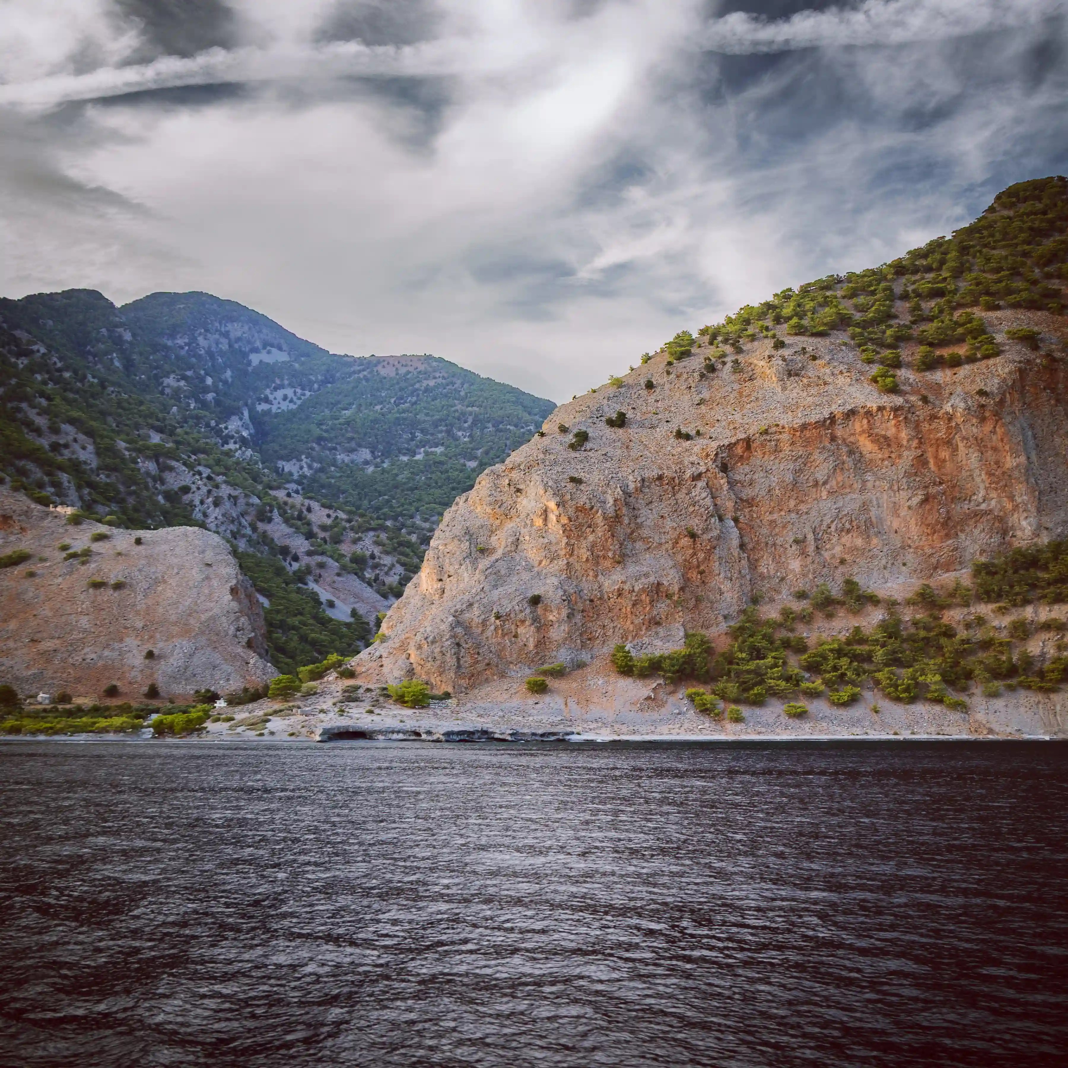 Rocky coastal cliffs rising above the sea with distant mountains under a cloudy sky.