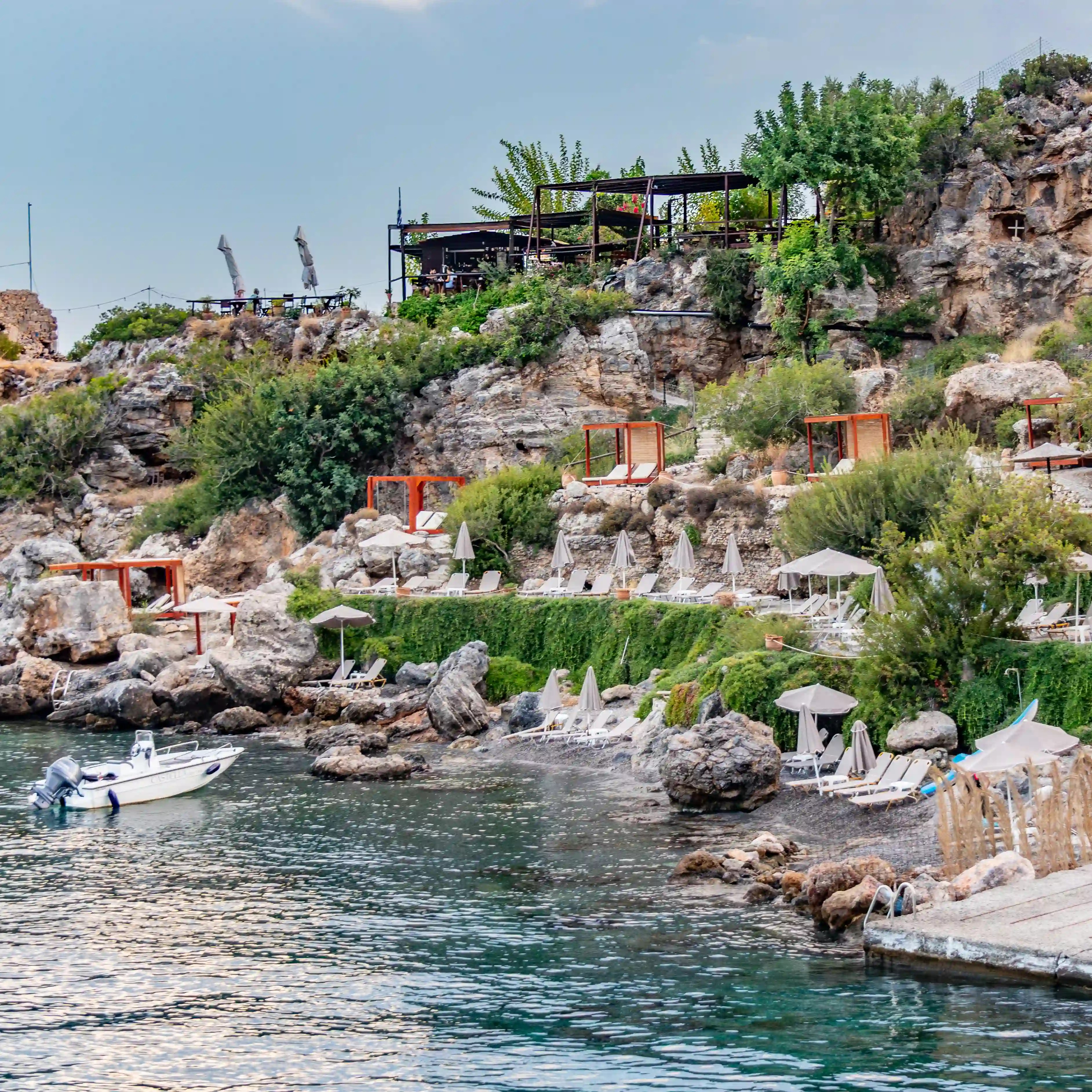 Rocky cove with sunbeds and umbrellas along the water and a café perched above the cliffs.