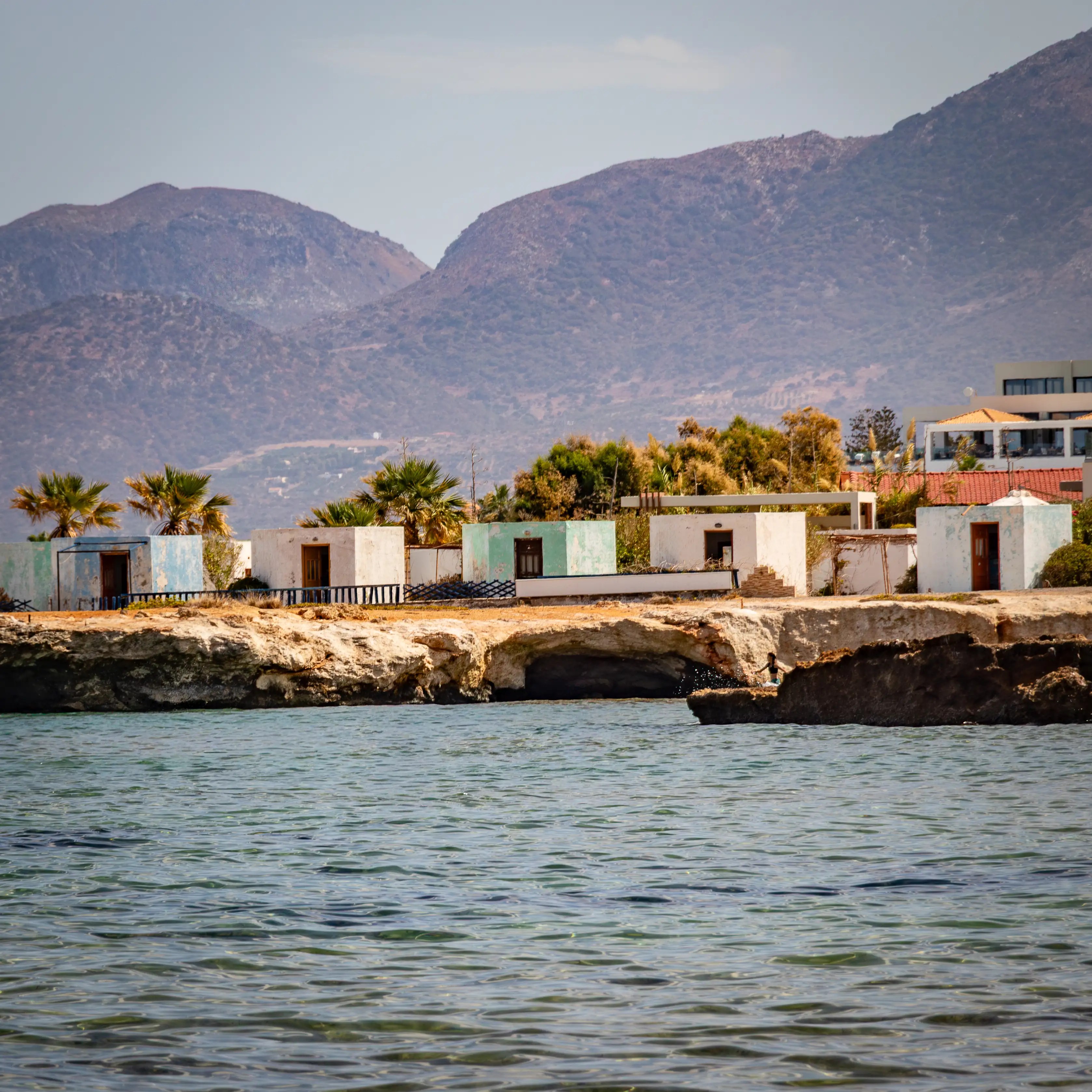 Small rustic seaside cottages on a rocky shoreline with the mountains of Crete in the background.