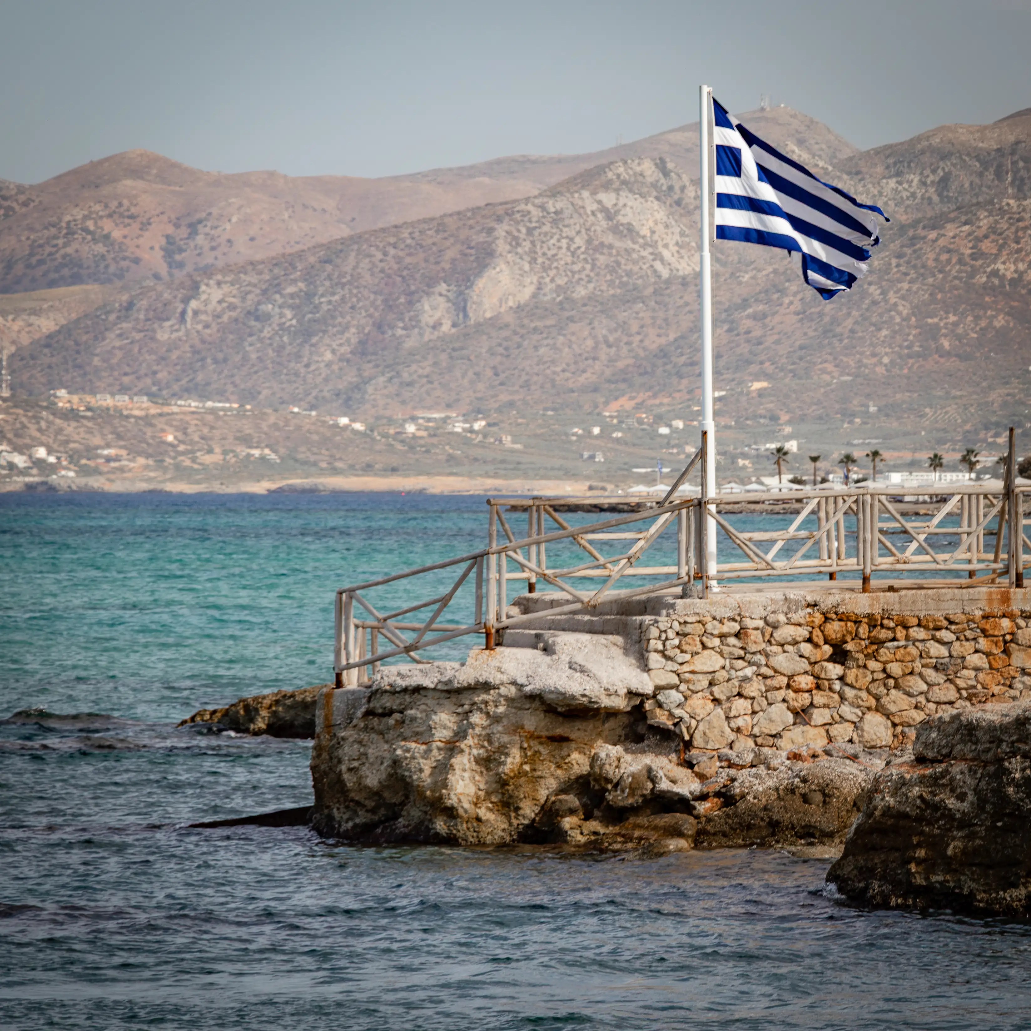Greek flag waving on a stone pier overlooking turquoise water with mountains in the background.