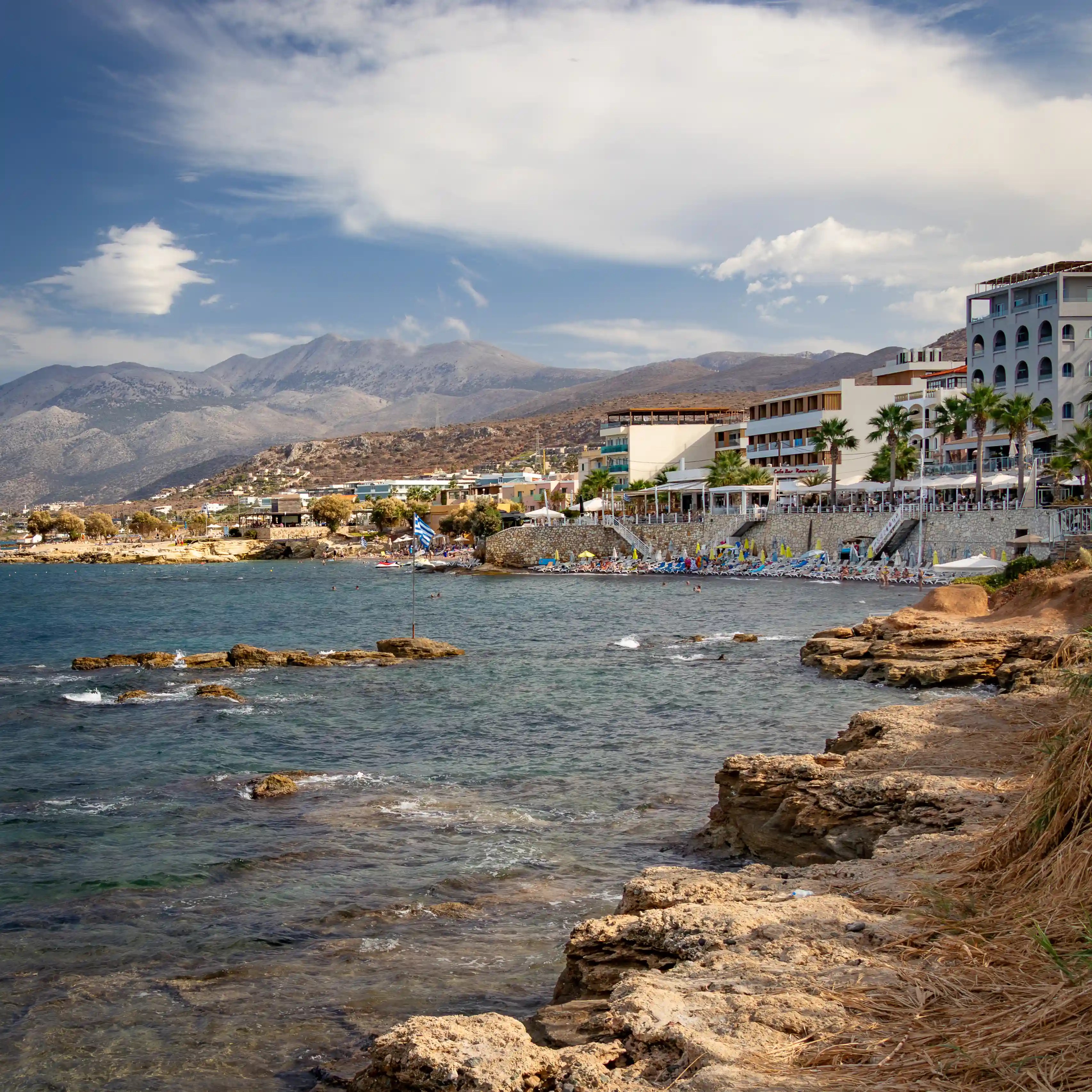 Rocky coastline with hotels and beachgoers along the Hersonissos shore and mountains in the distance.