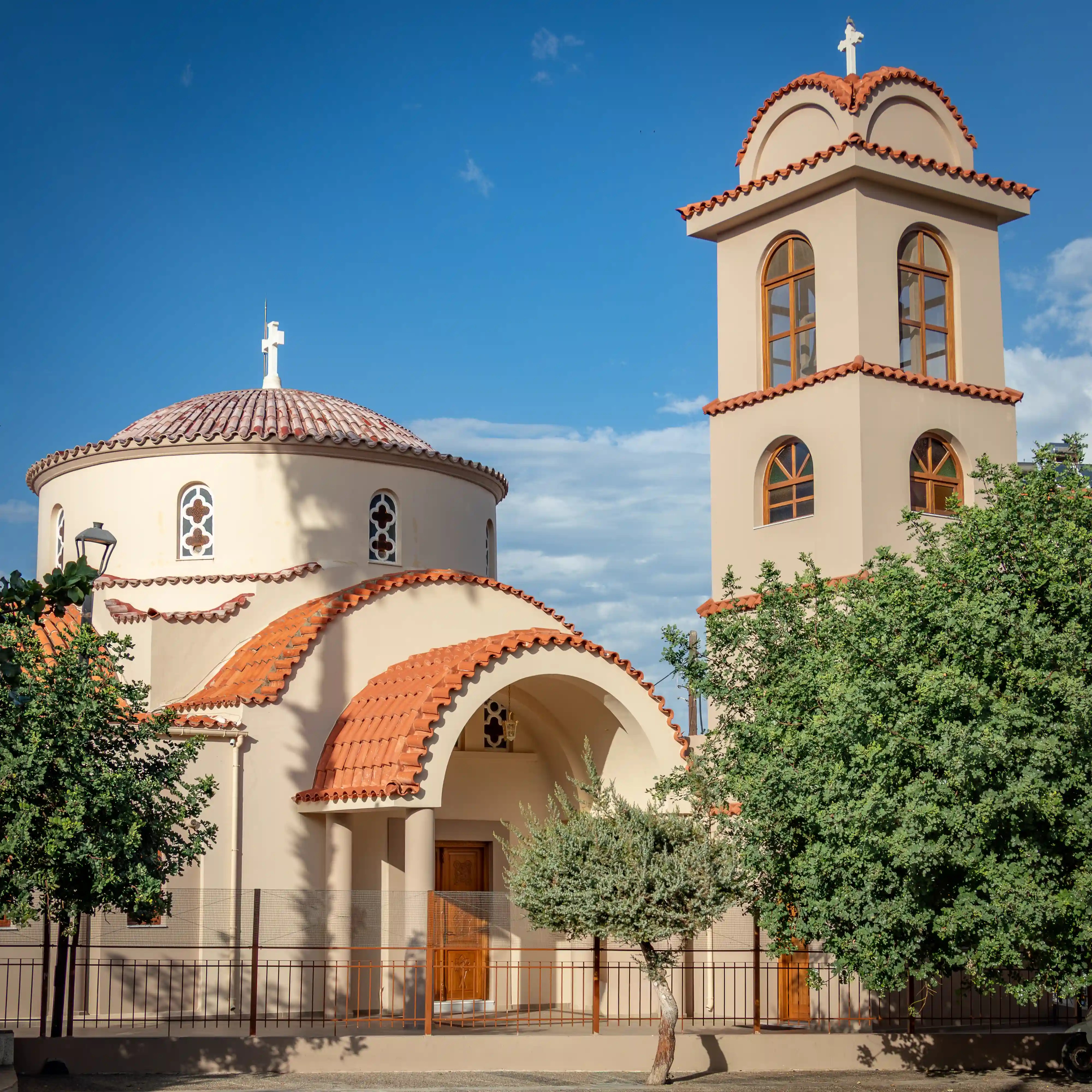 Cream-colored Greek Orthodox church in Hersonissos with a tiled dome and bell tower against a blue sky.