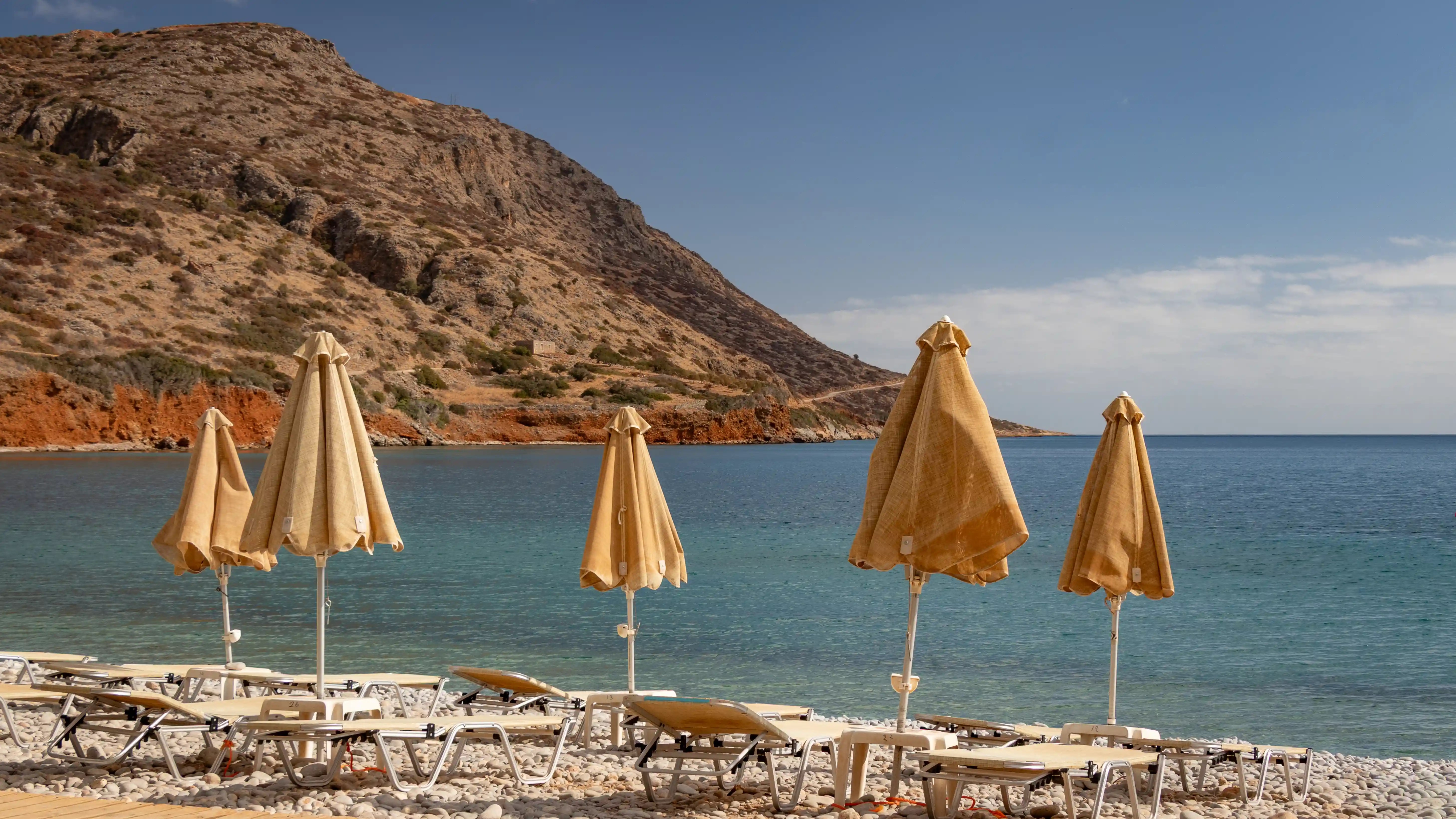 Closed beige beach umbrellas and sun loungers line a pebble beach in Plaka, with a rocky hillside rising beside the clear blue water.