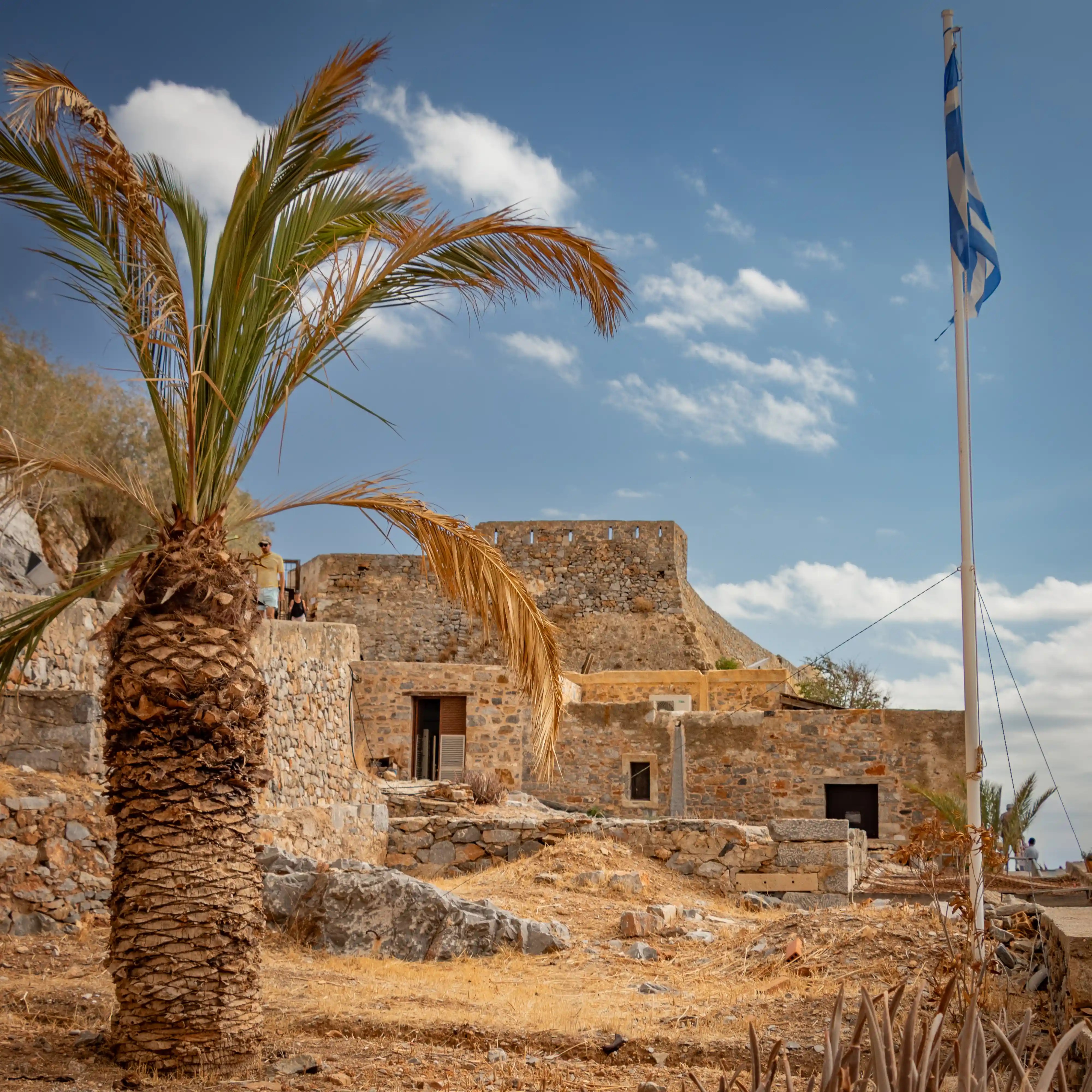 A palm tree stands in front of Spinalonga’s stone buildings with a Greek flag flying against a blue sky.