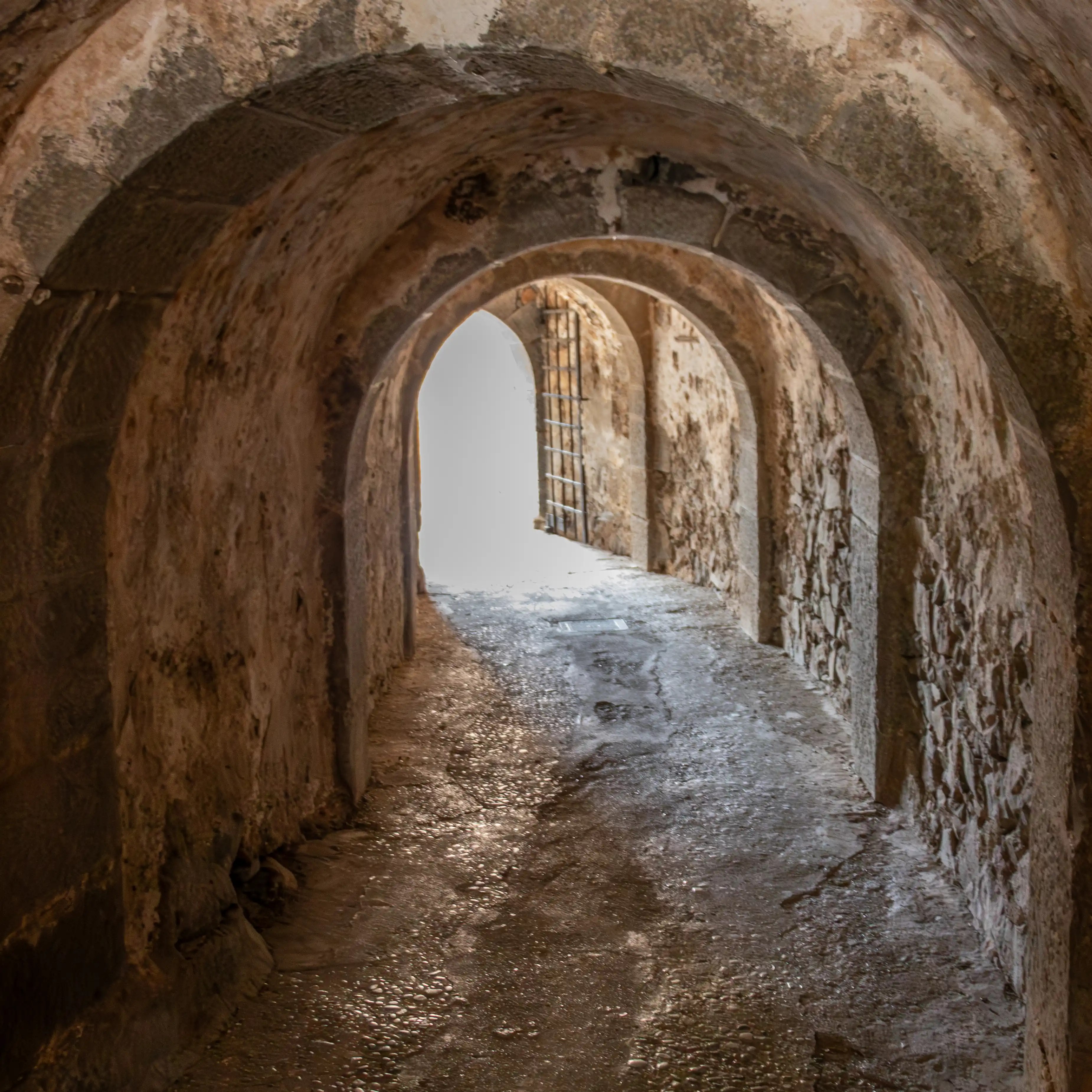 A stone tunnel on Spinalonga Island leads toward a bright opening framed by an old iron gate.