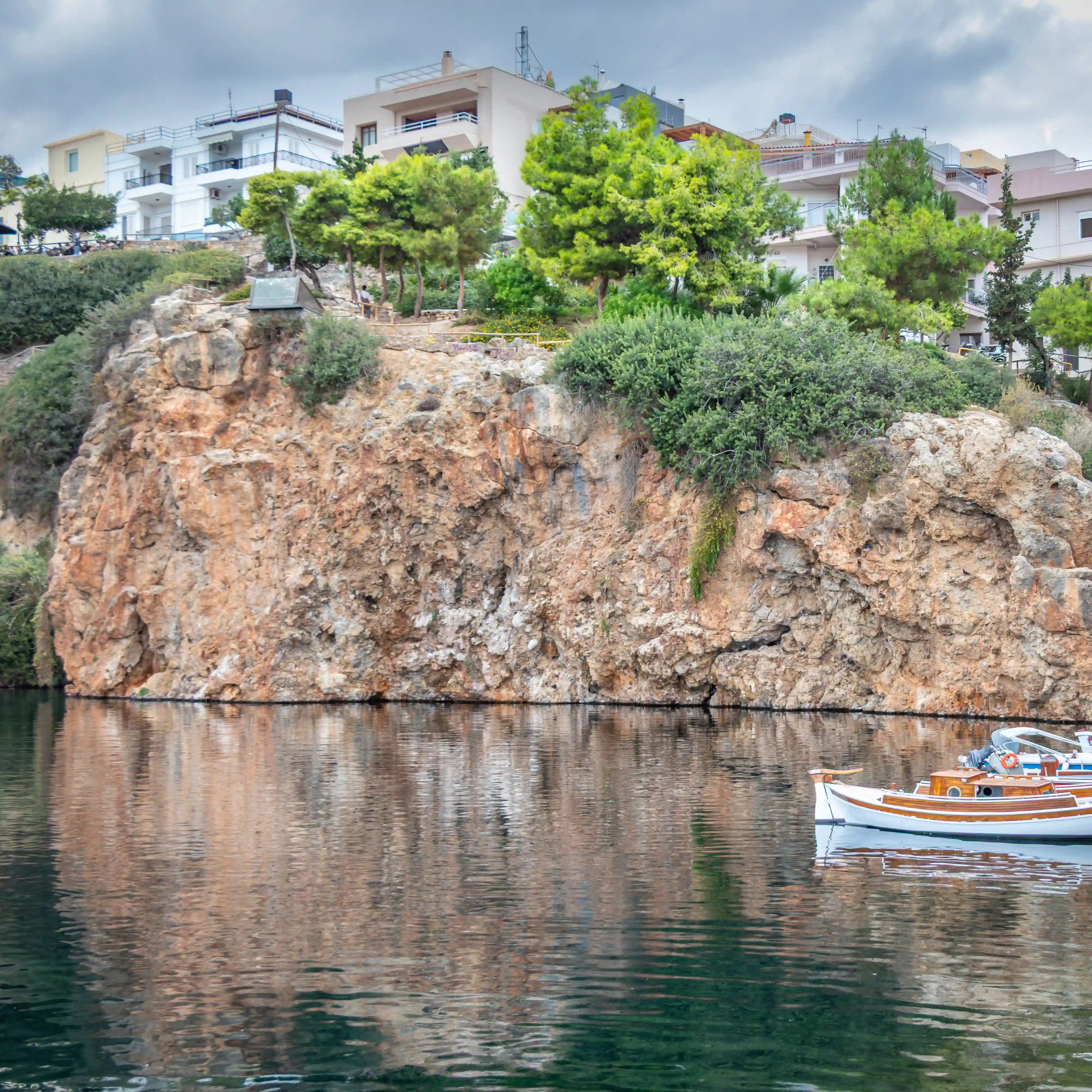 A rocky cliffside topped with homes and trees reflected in the still water below, with a small wooden boat on the right.