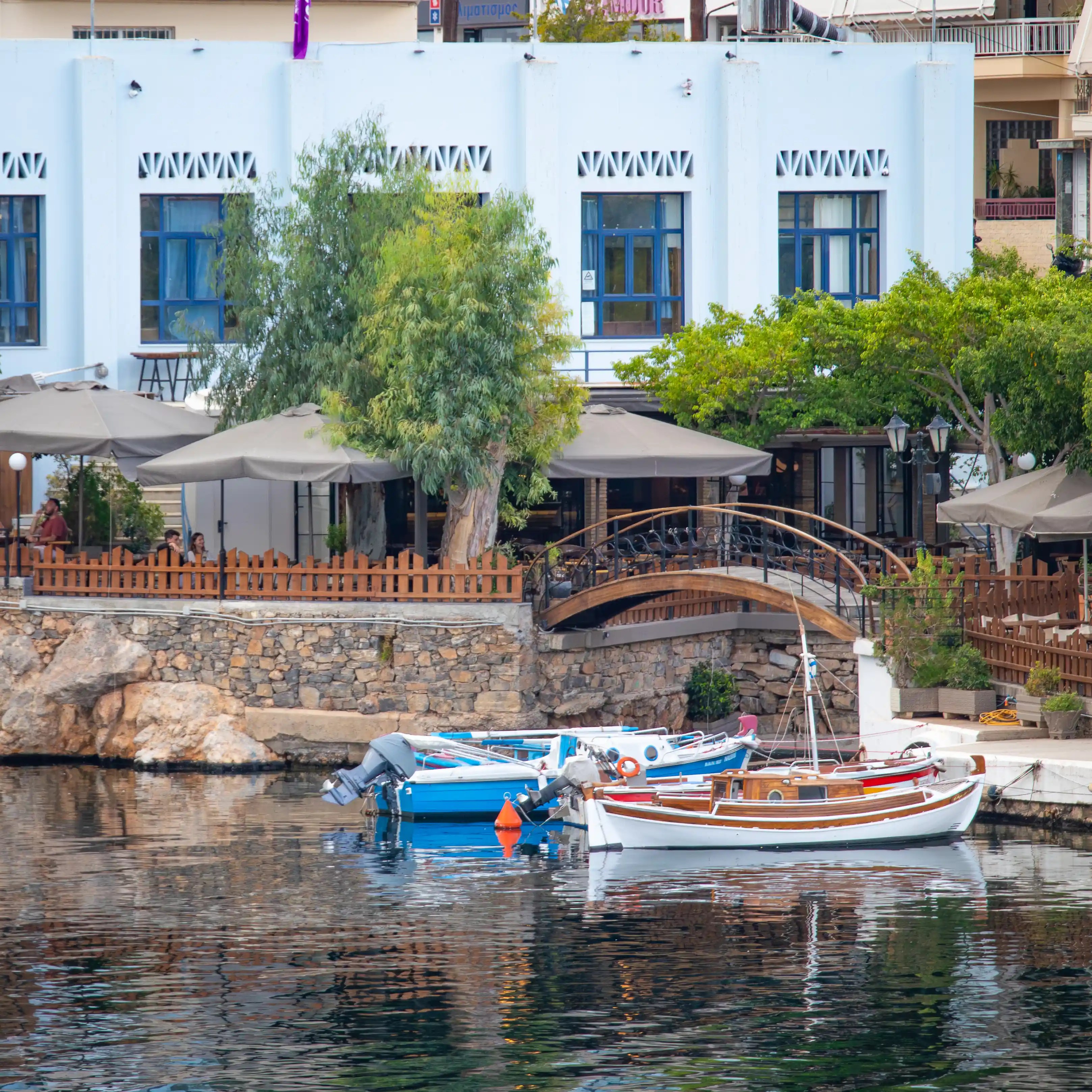 Small boats floating on calm water near a waterfront café with umbrellas and trees.