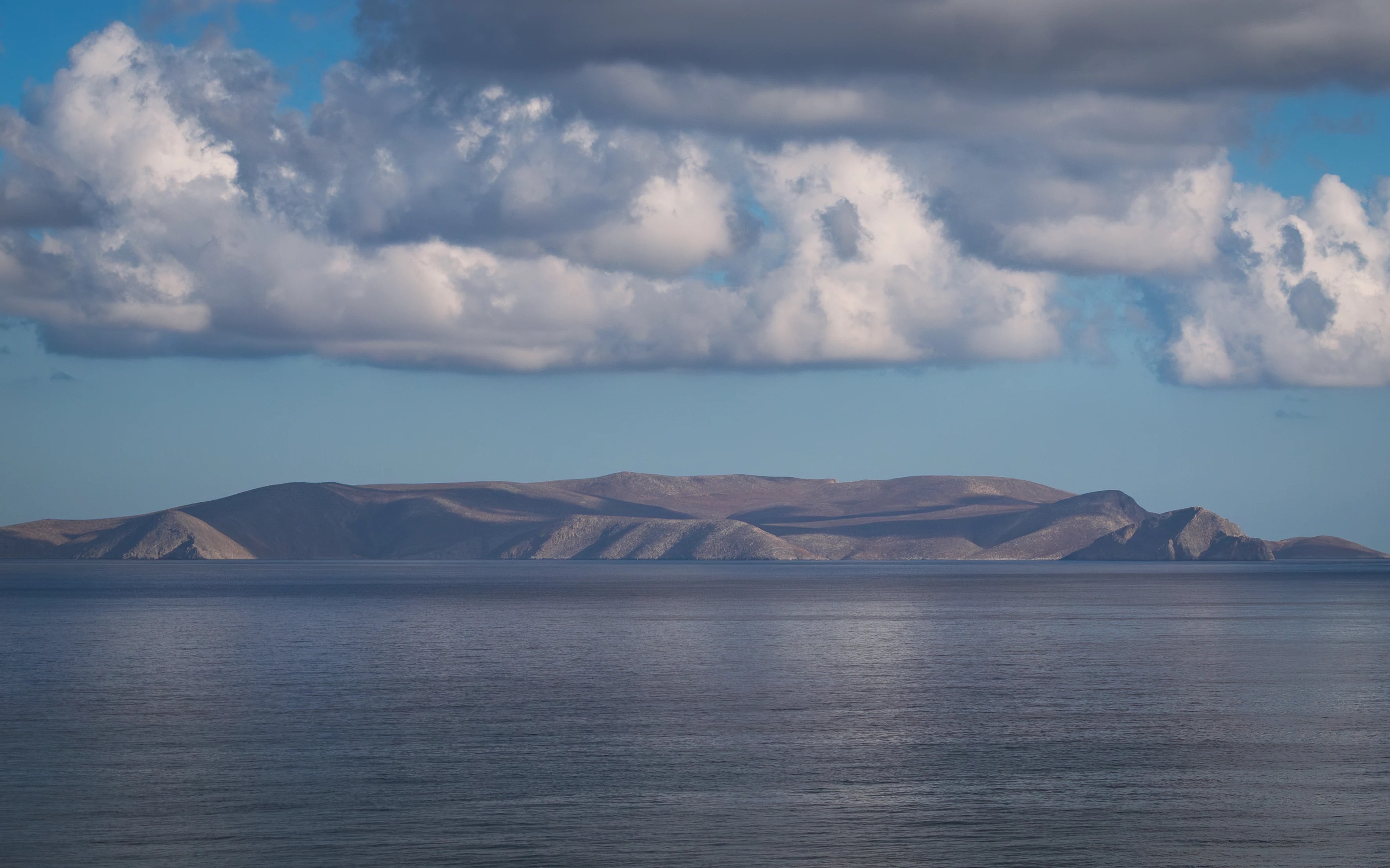 A distant view of Dia Island with its long, rocky ridgeline beneath large, dramatic clouds over the blue Sea of Crete.