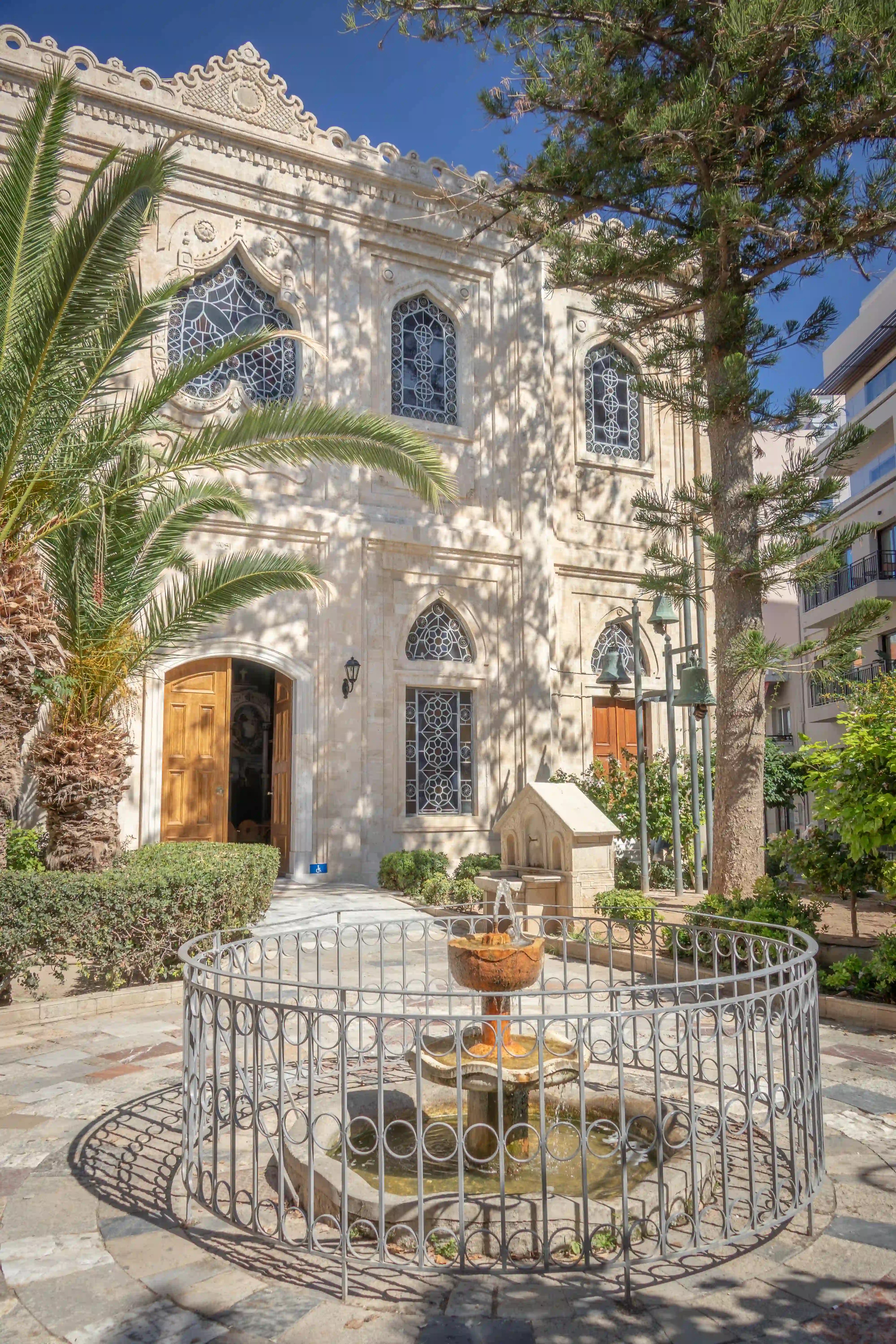 Stone Agios Titos Church façade with pointed windows, palm trees, and a small courtyard fountain.