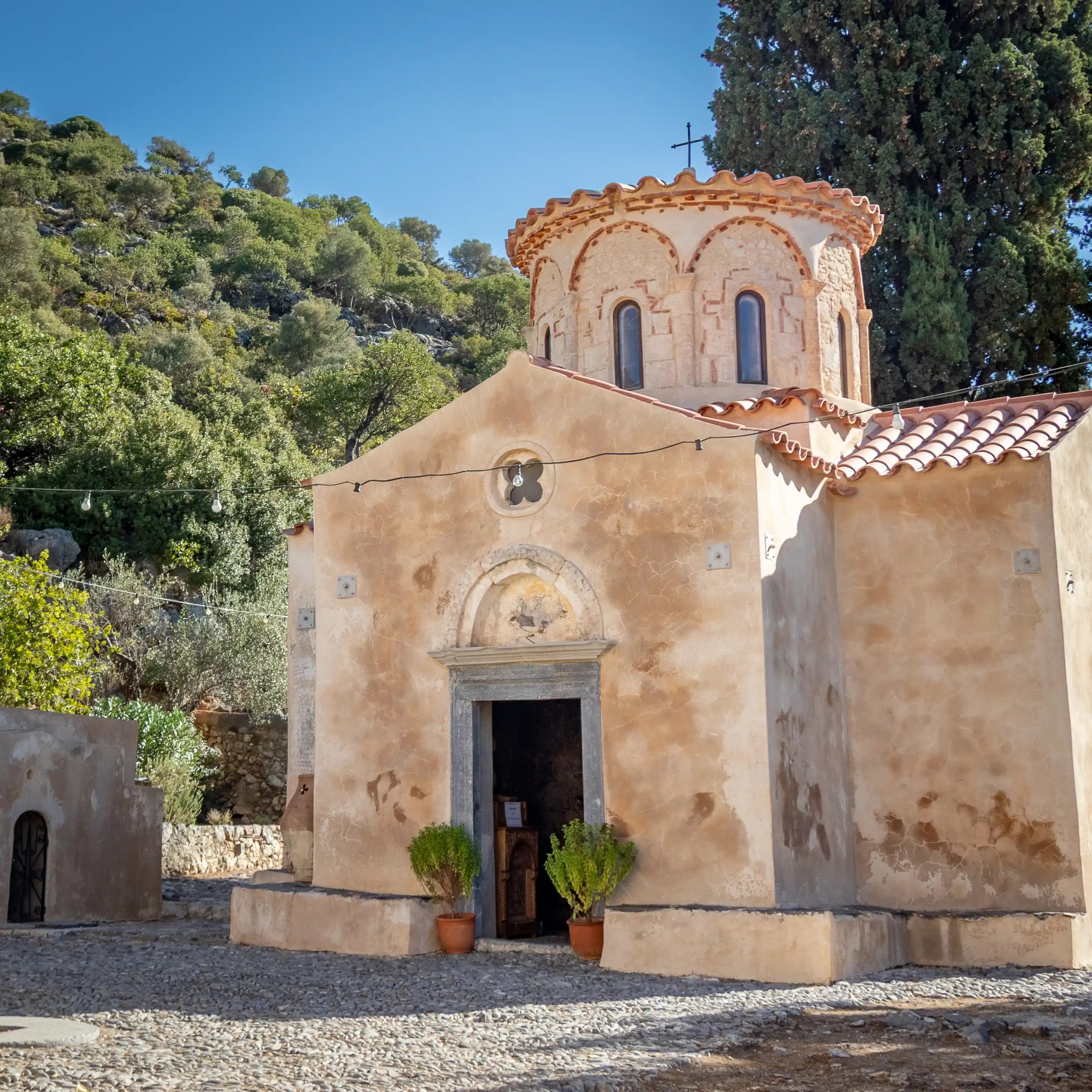 Front view of Panagia Gouverniotissa Monastery, a small Byzantine church with a rounded dome, pale stone walls, and terracotta roof tiles set against a green hillside.
