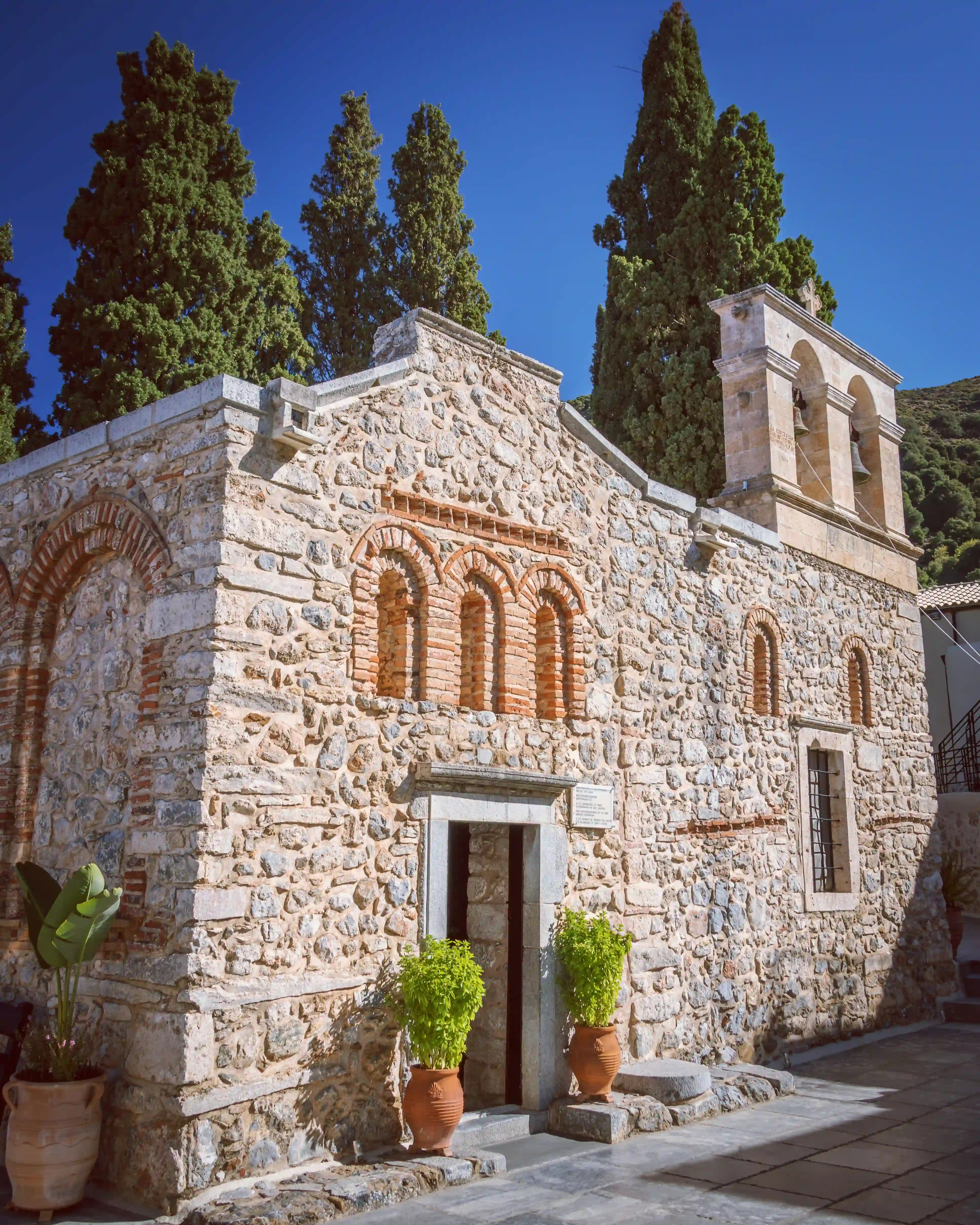 Stone exterior of the Monastery of the Panayia Kera with terracotta accents, narrow windows, and tall cypress trees behind it.