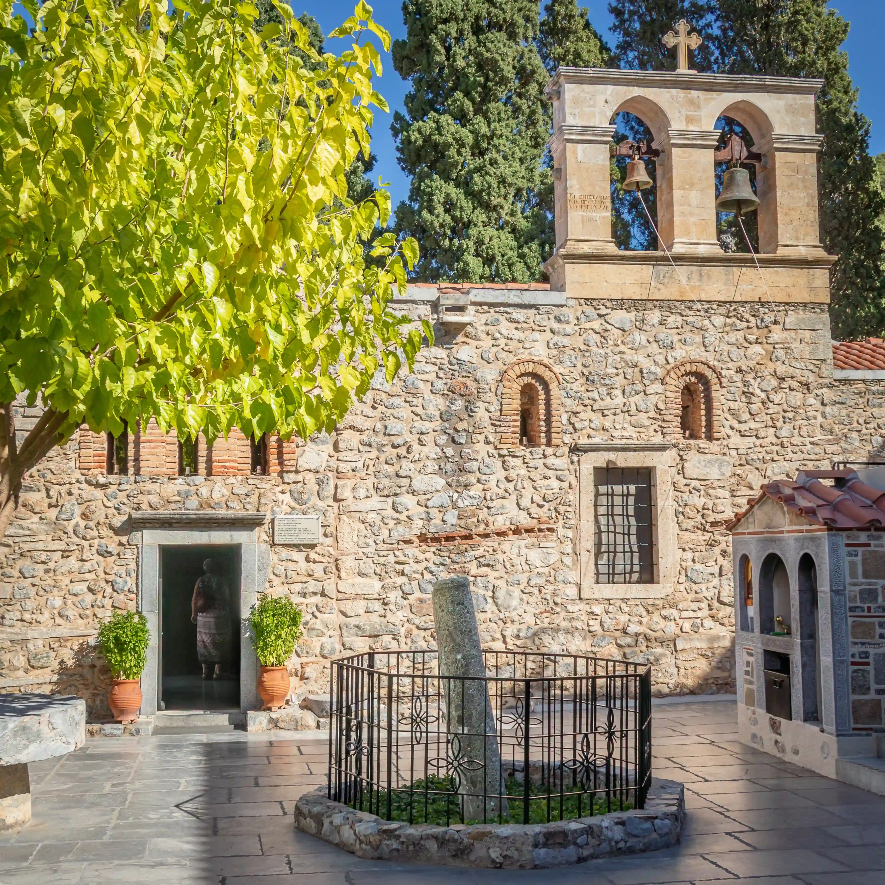 Stone courtyard of Kera Kardiotissa Monastery with bell tower, leafy tree, and fenced marble column in the center.