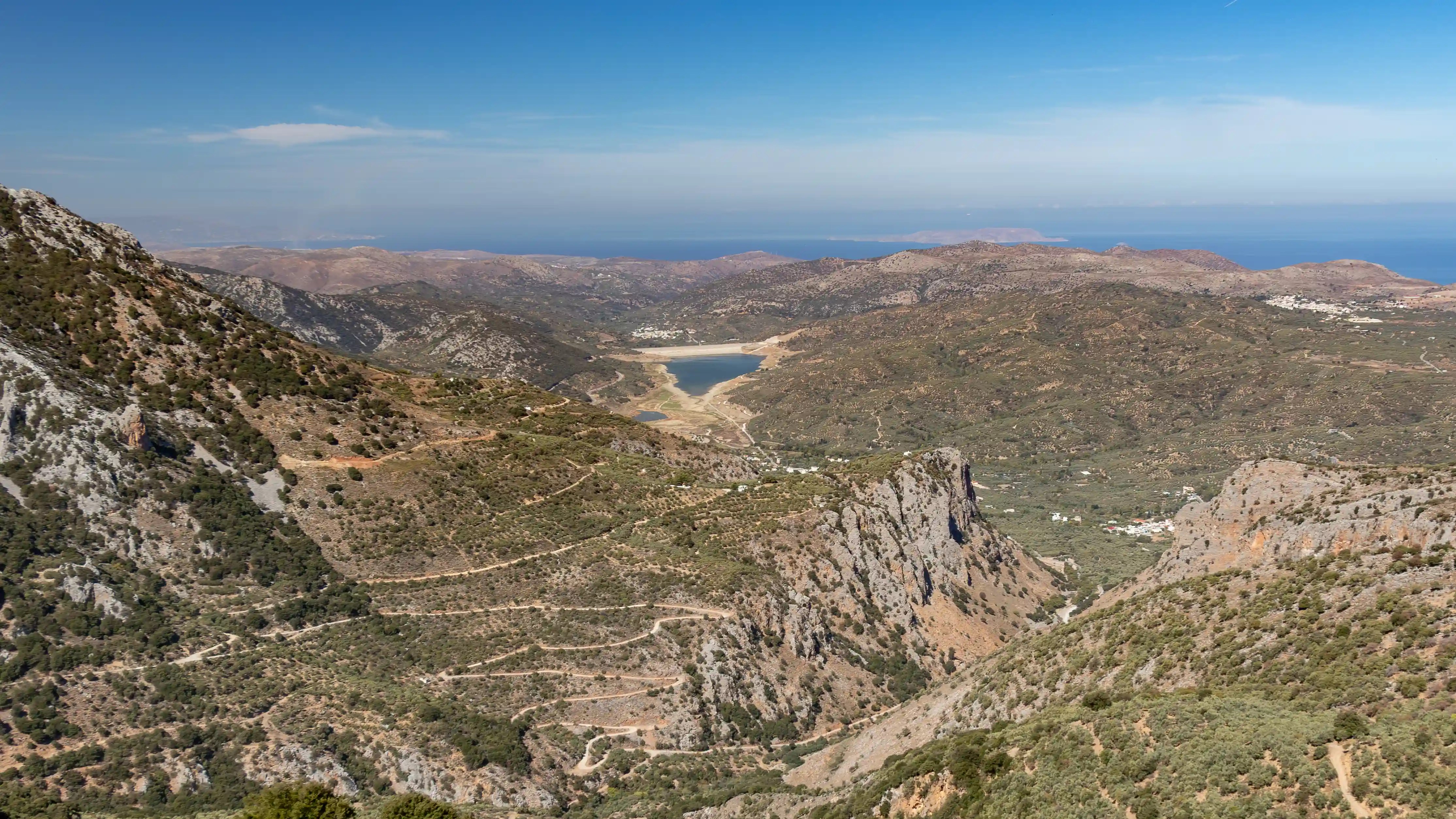 Wide view over mountain slopes, winding roads, and a small reservoir in the Lasithi Plateau region of Crete.