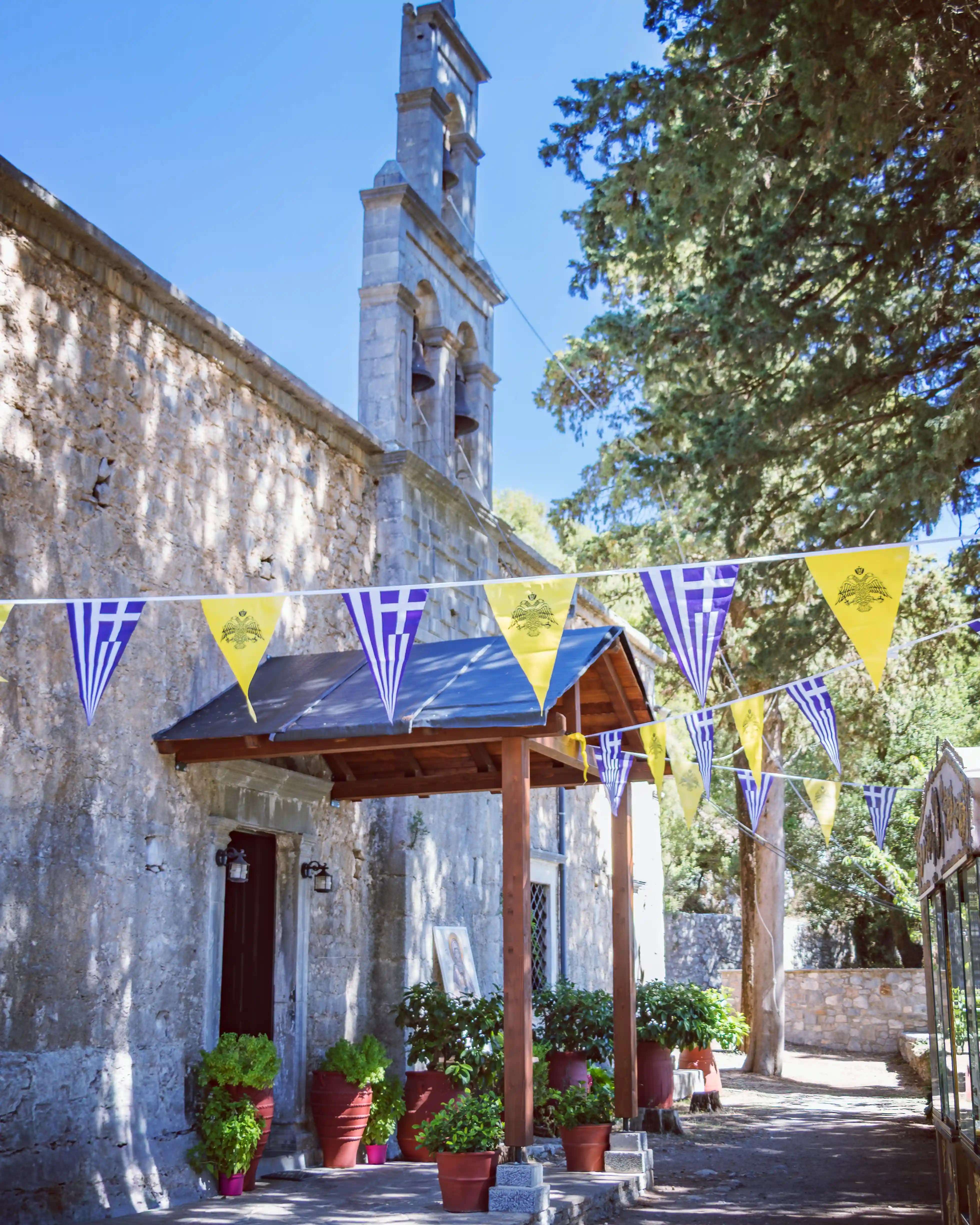 Entrance of the Holy Monastery of Vidiani with stone walls, yellow and blue Orthodox flags, potted plants, and a tall bell tower rising above the trees.