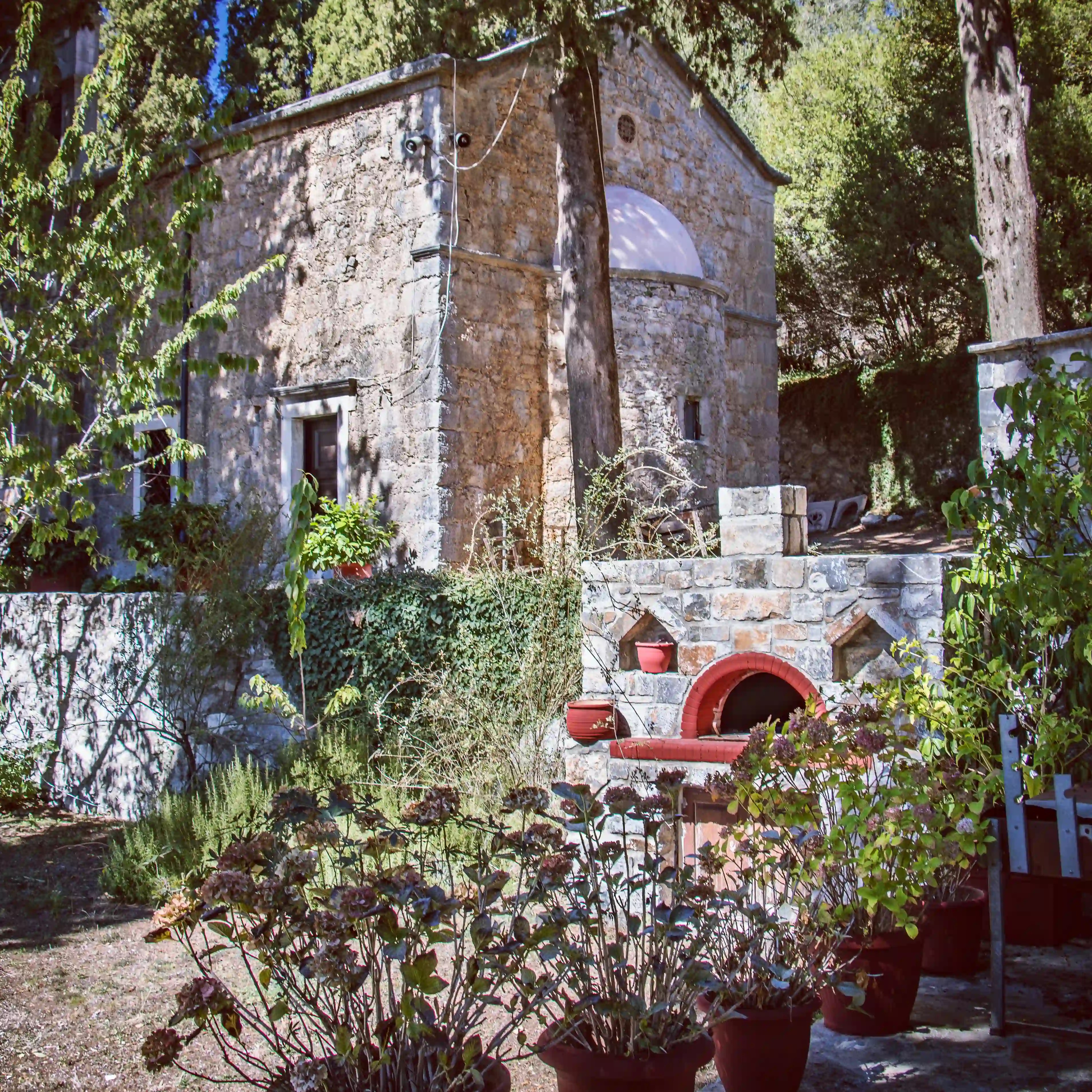 Exterior view of the stone chapel at the Holy Monastery of Vidiani surrounded by trees and garden plants.