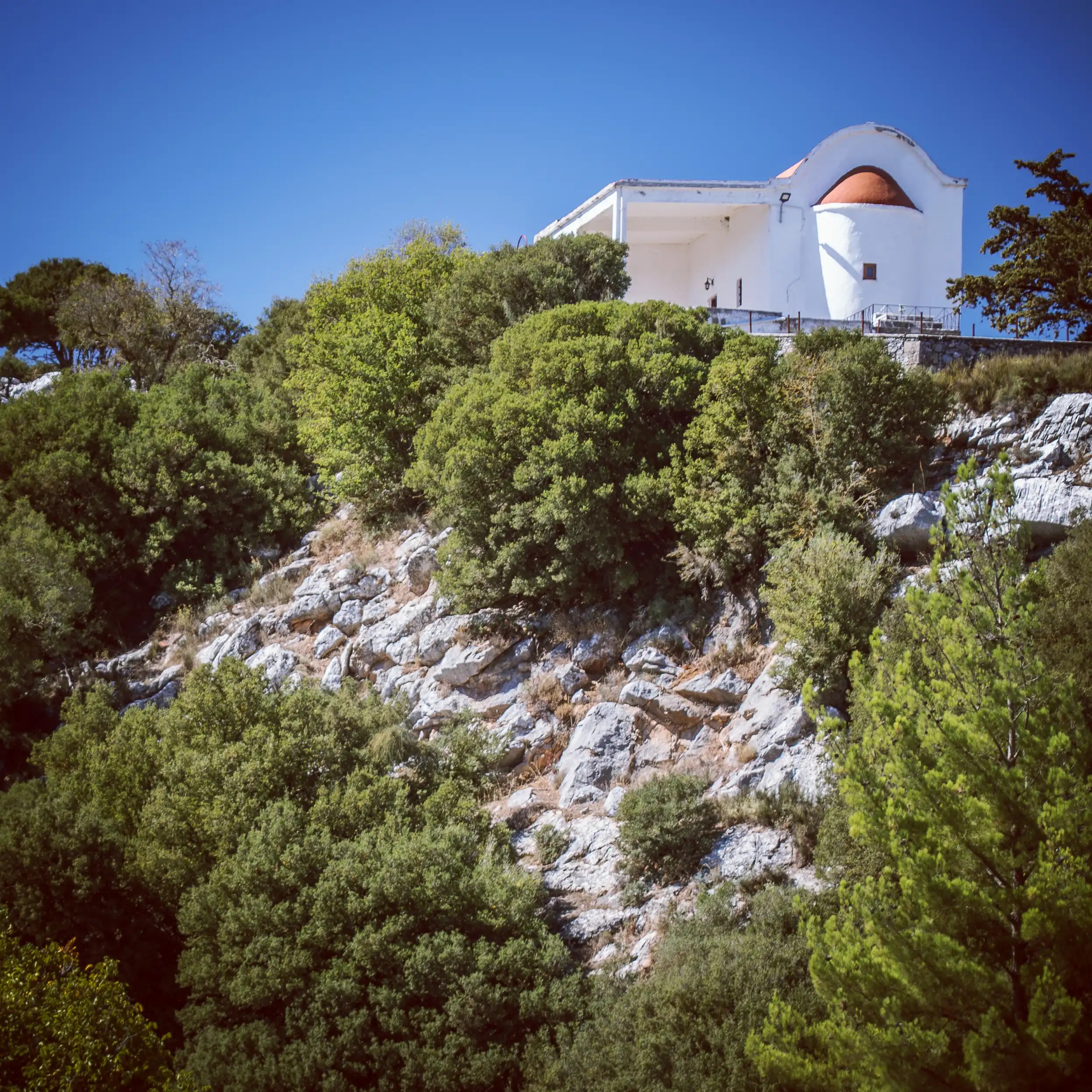 Small white chapel with an orange dome perched on a rocky hillside surrounded by trees.
