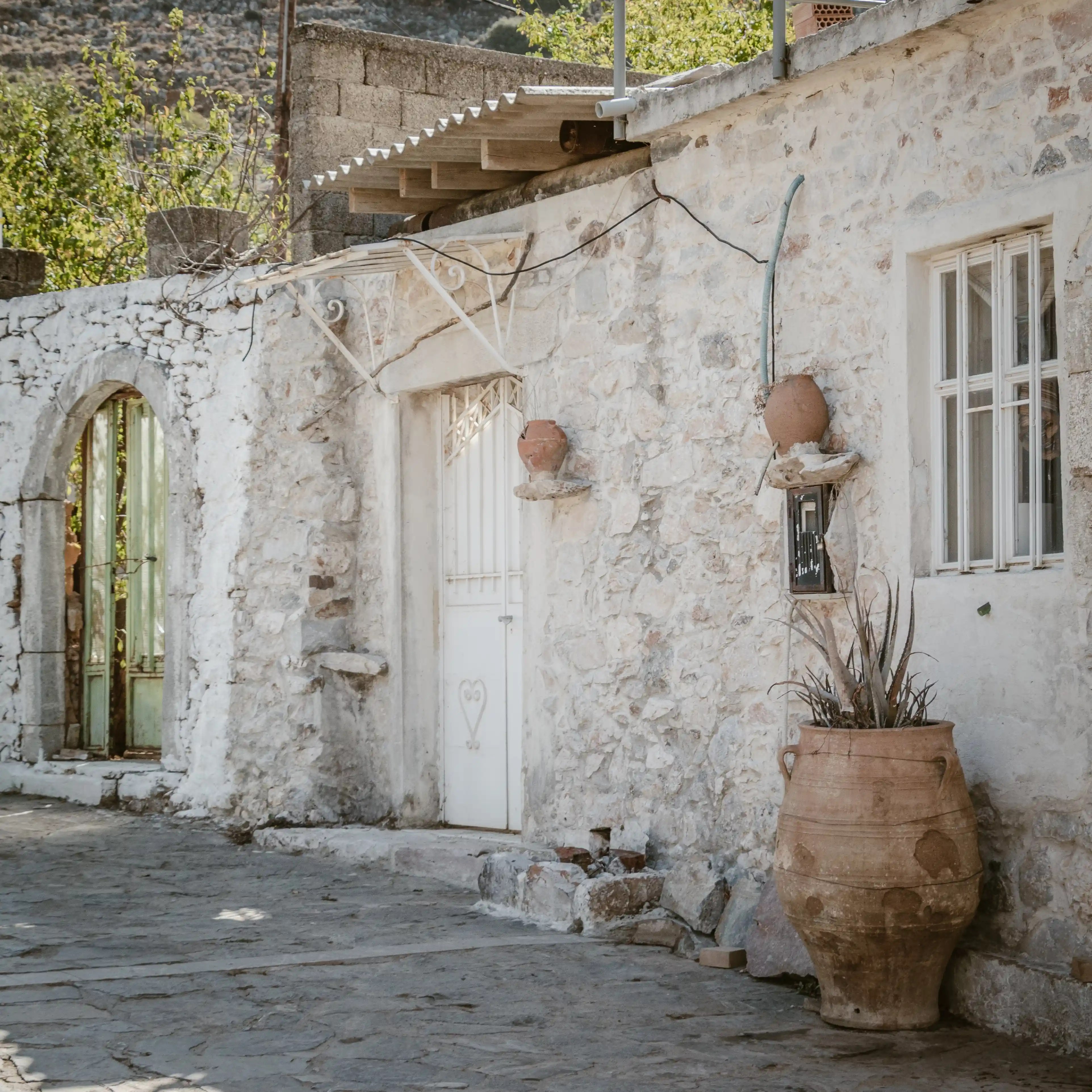 Stone house in Krasi with a white door, clay pots, and a large terracotta jar beside the wall.