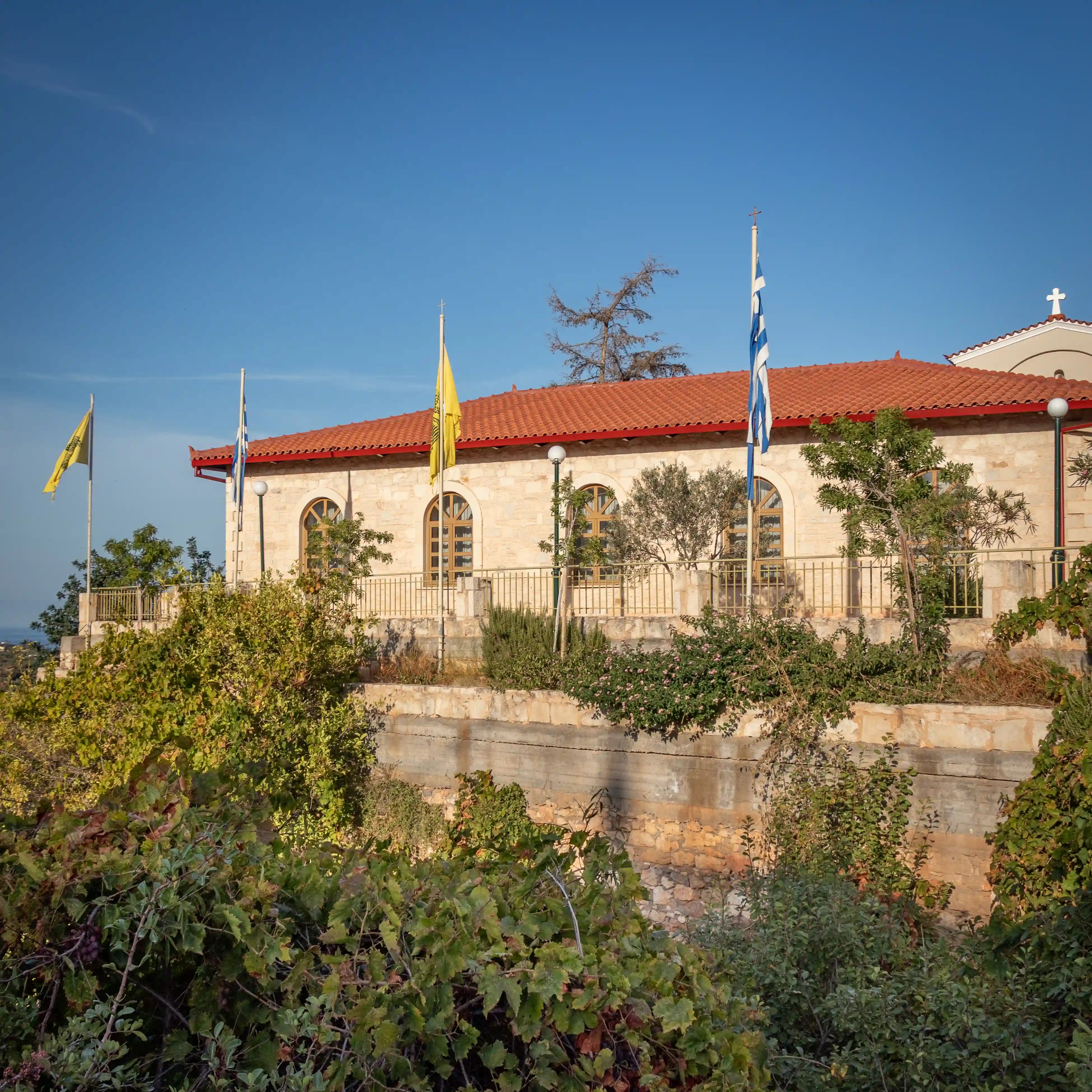 Stone church on a hillside with Greek and ecclesiastical flags.
