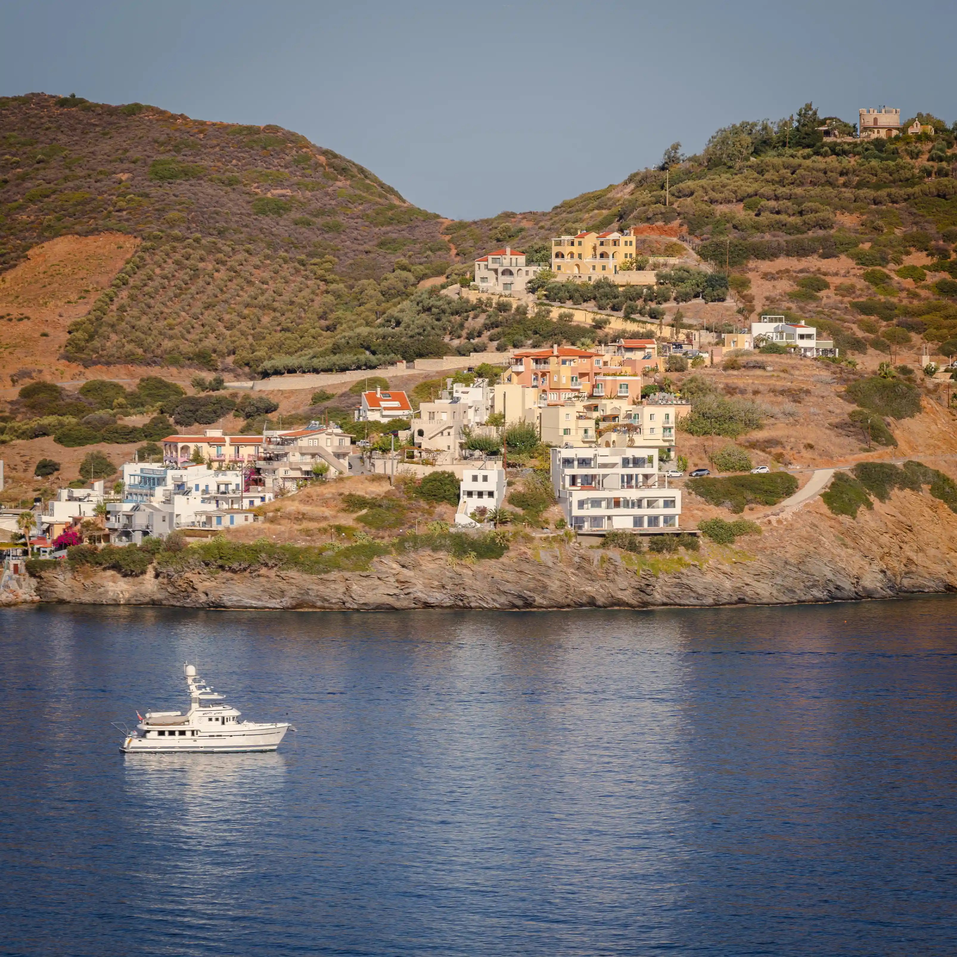 Coastal hillside village with colorful homes above the sea and a small yacht below.
