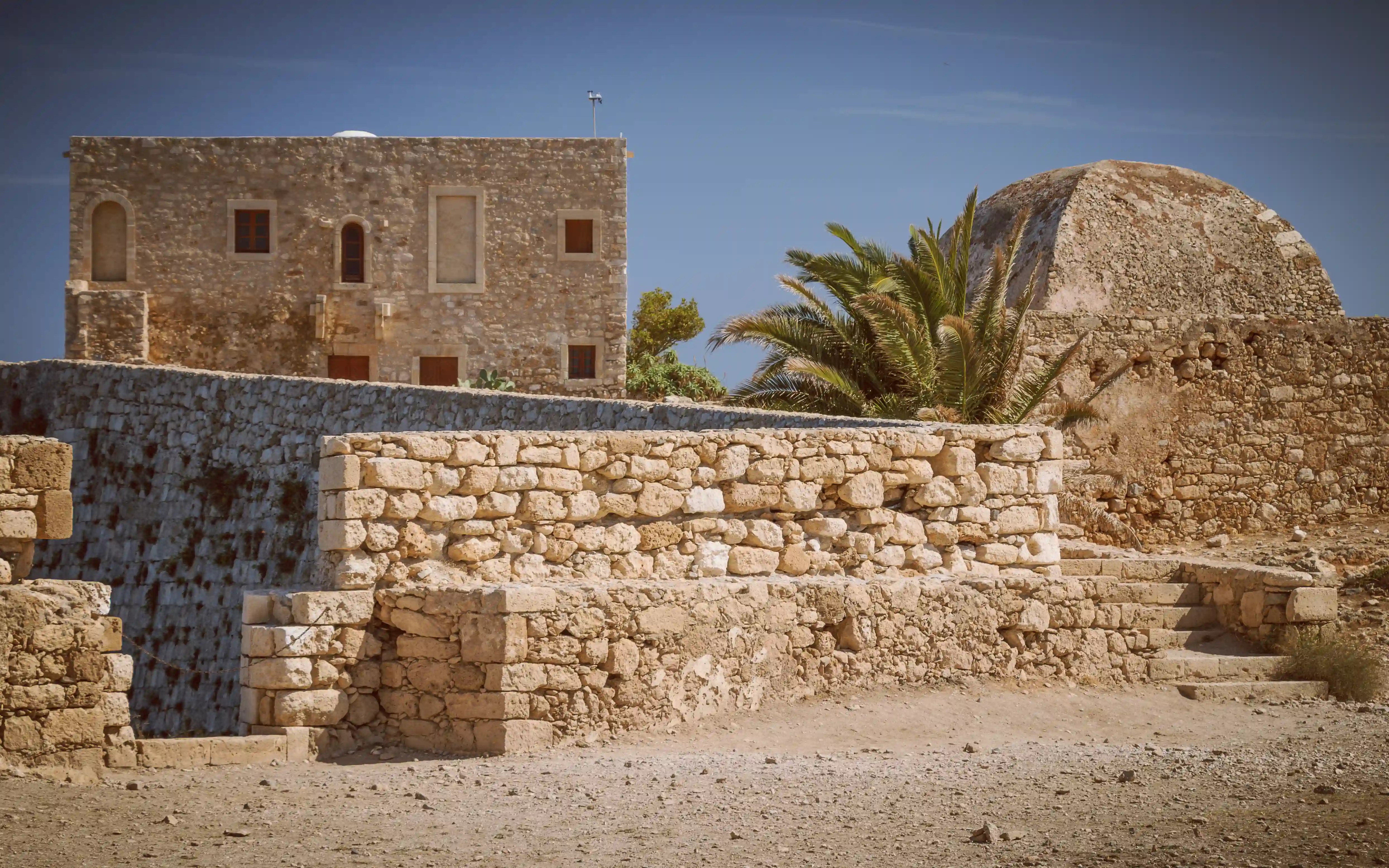 Sunlit stone buildings and walls inside the Rethymno Fortezza with a large domed roof visible behind them.
